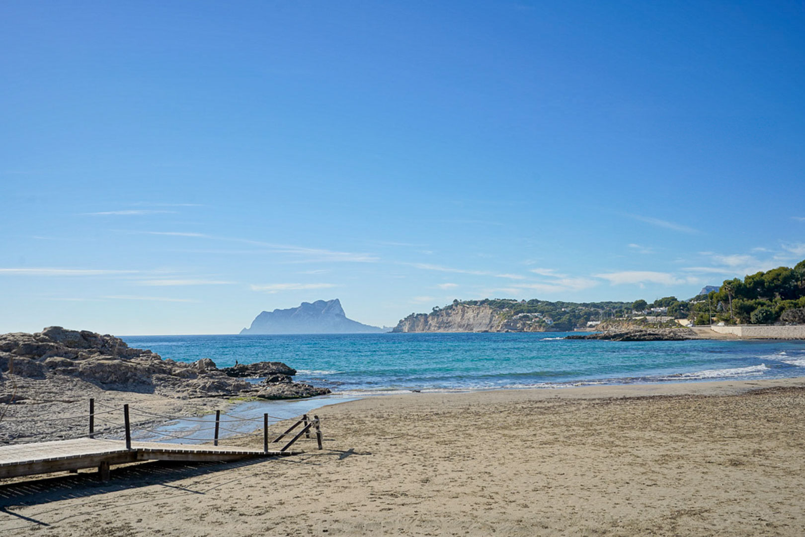 Coastal view in Moraira featuring a wooden beach access ramp and the iconic Peñón de Ifach rock formation visible across the Mediterranean horizon.