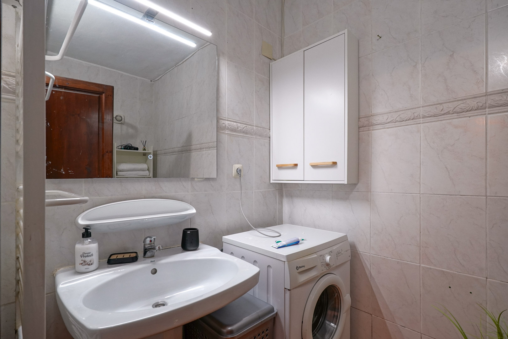Functional bathroom featuring a ceramic sink, LED-lit mirror, and an integrated laundry area with a washing machine and wall-mounted storage cabinet.
