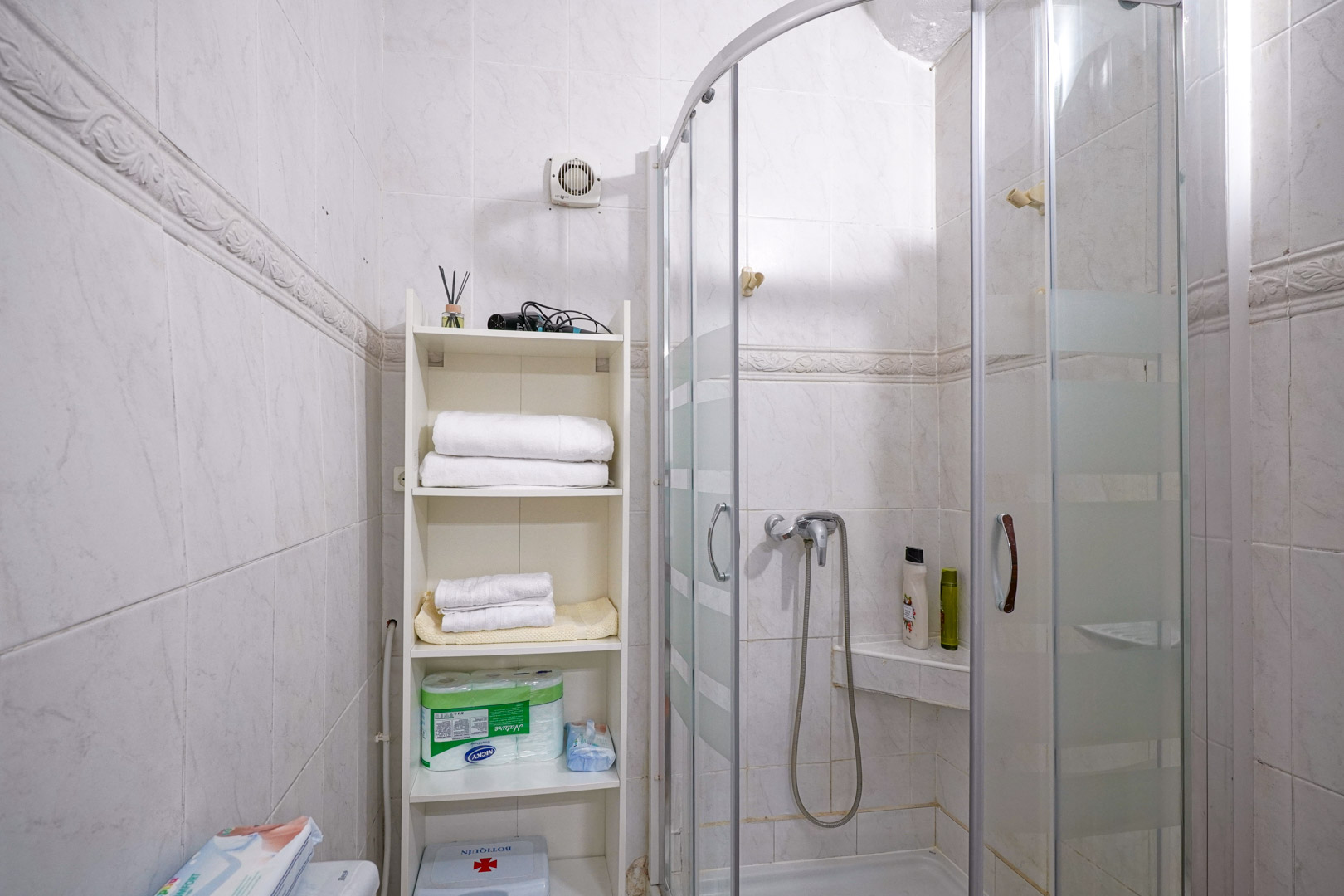 Functional bathroom featuring a curved glass shower enclosure and white tiled walls with a decorative border. Includes a white shelving unit for practical storage.