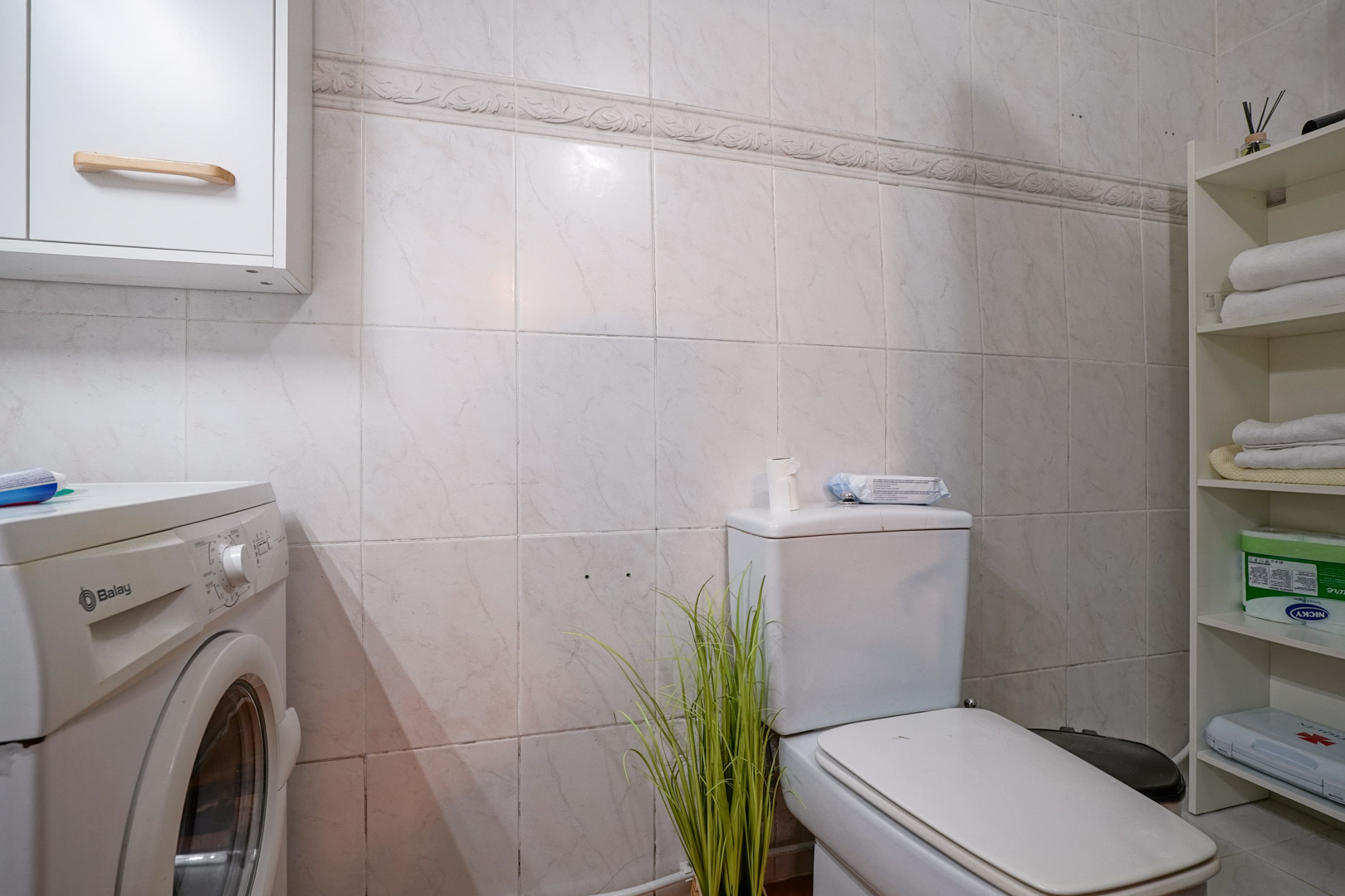 Functional bathroom with laundry area, featuring a Balay washing machine, storage shelving, and tiled walls with a decorative border.