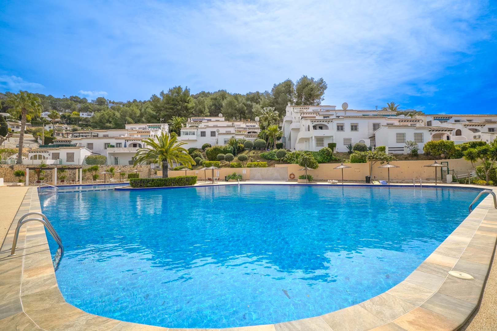 Large communal swimming pool with a curved design, featuring stone tiling, straw parasols, and landscaped gardens set against the Mediterranean architecture of Moraira.
