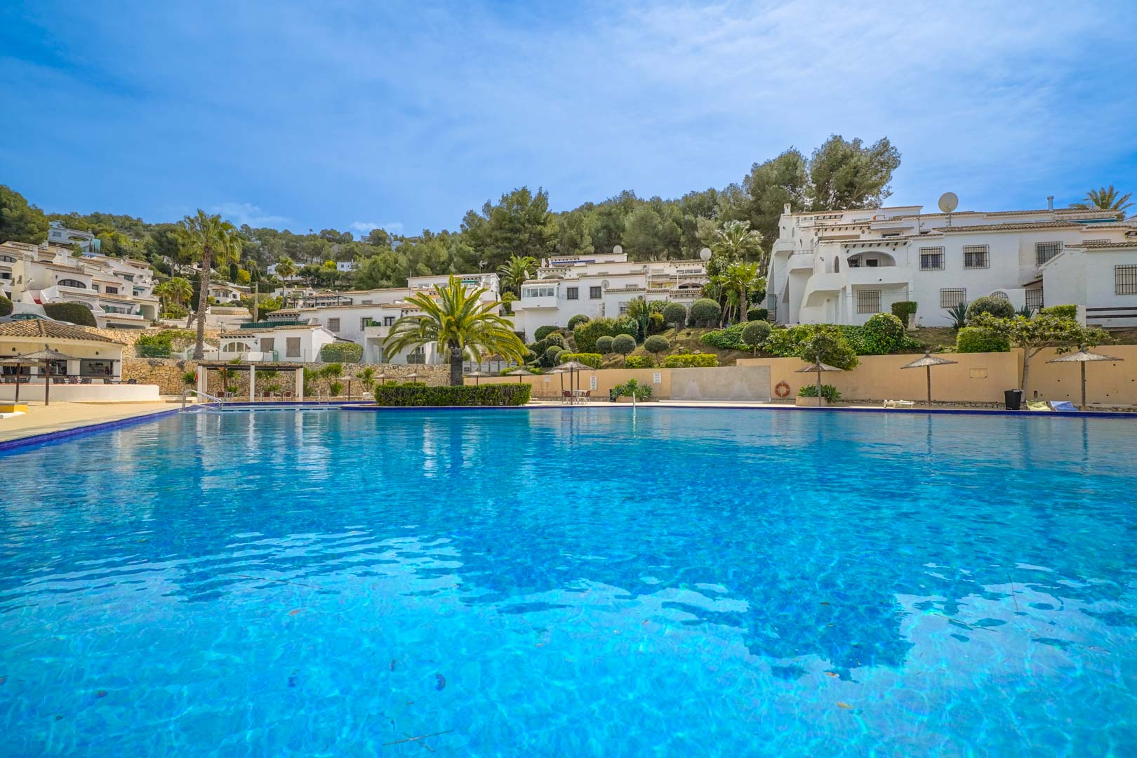 Large communal swimming pool with blue tiling, surrounded by Mediterranean landscaping and white-washed buildings set against a clear sky in Moraira.