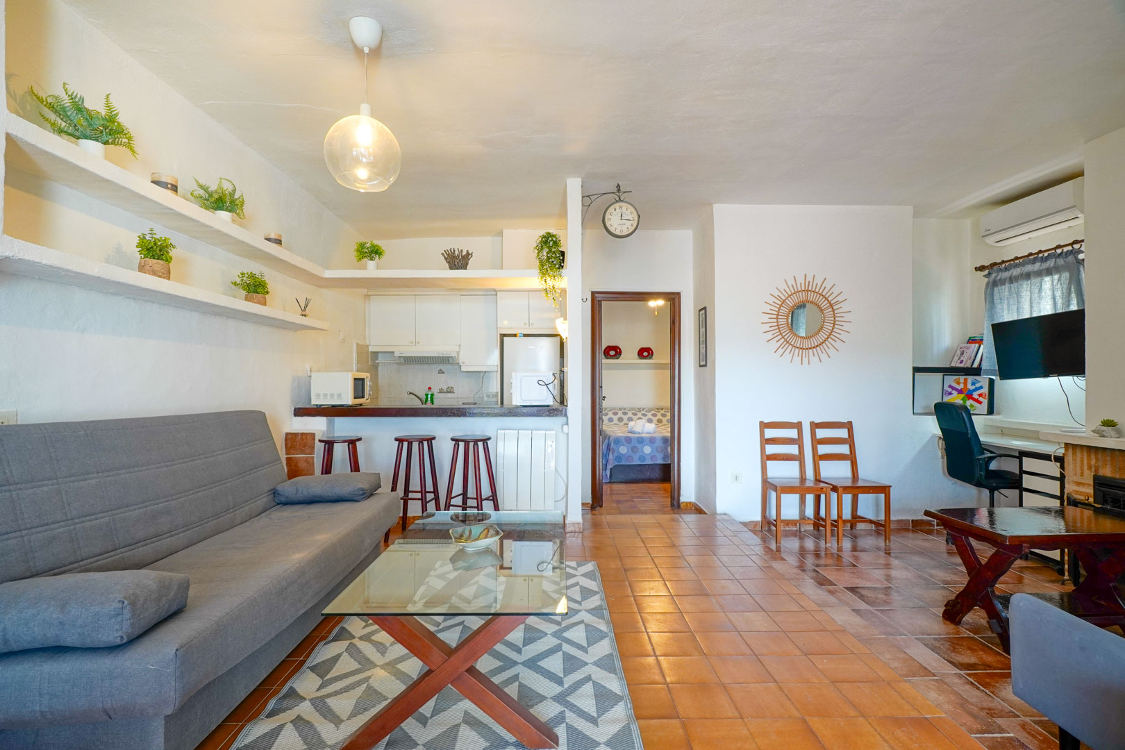 Open-plan living area featuring a semi-integrated kitchen with breakfast bar, rustic tile flooring, and built-in shelving. The layout includes a workspace and access to the bedroom.