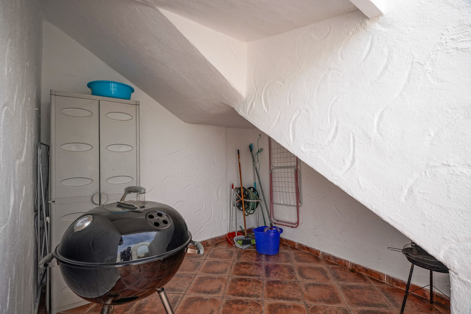 Outdoor storage area under the staircase featuring terracotta tile flooring, a resin cabinet, and space for a kettle grill and maintenance tools.