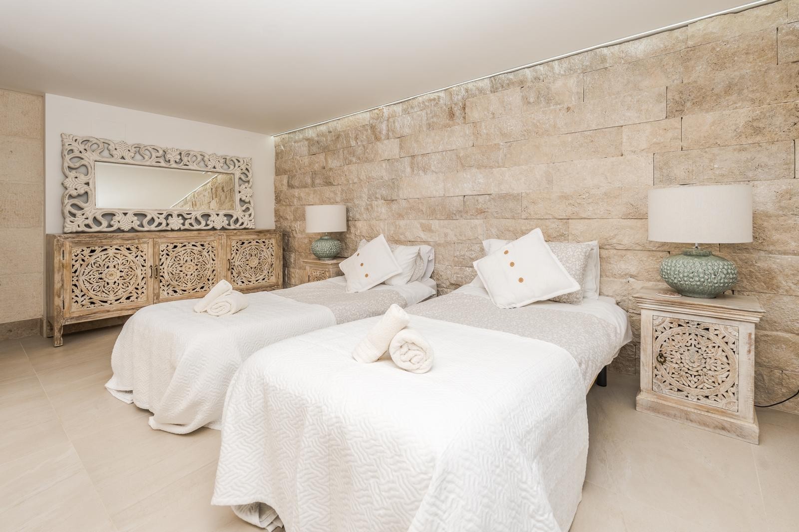 Bedroom featuring a natural stone accent wall, twin beds, and intricate hand-carved wooden furniture. The space is finished with neutral tones and soft ambient lighting.