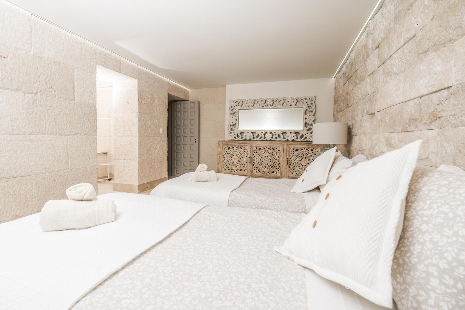 Bedroom featuring natural stone walls, twin beds, and intricately carved wooden furniture. Perimeter LED lighting highlights the textured masonry and elegant decor.
