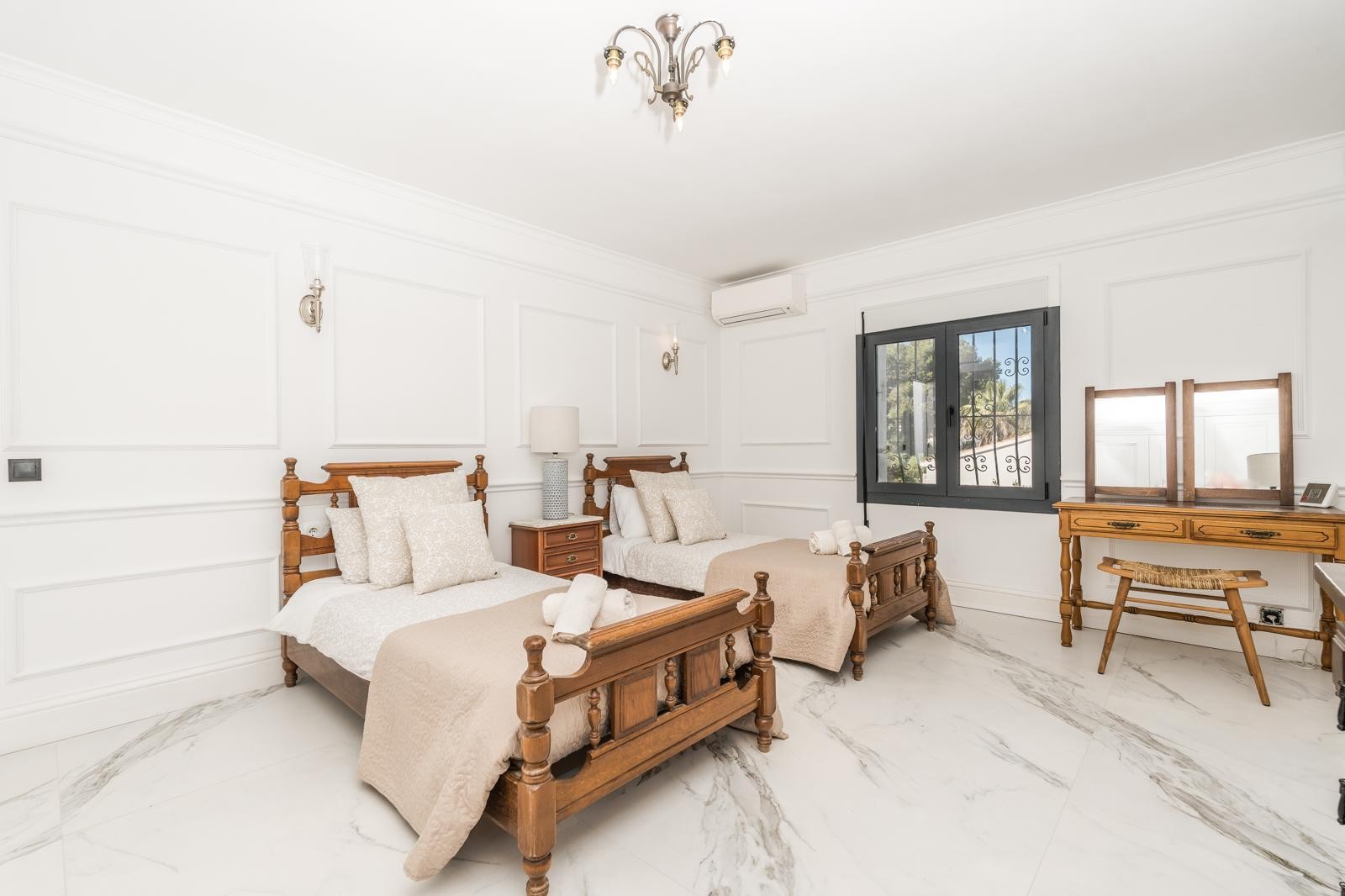 Bright bedroom featuring twin wooden beds, marble flooring with grey veining, and classic wall moldings. Includes a wooden vanity desk and split-system air conditioning.