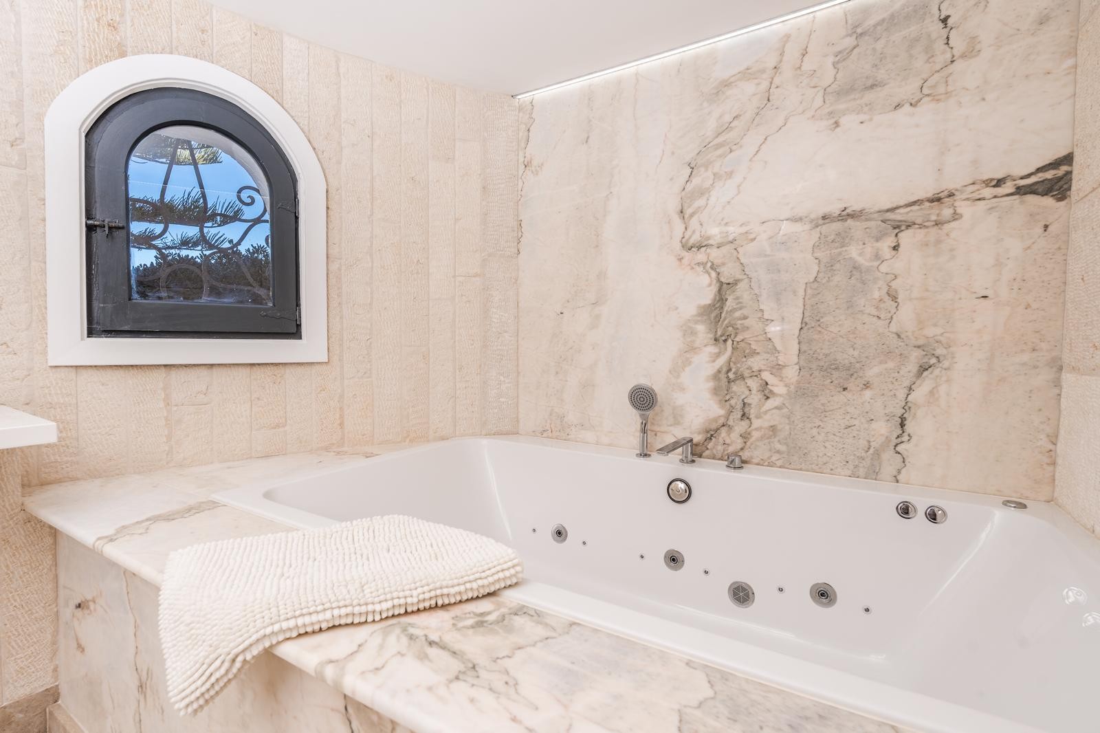 Built-in whirlpool bathtub featuring elegant marble surfaces and textured stone walls. A classic arched window and recessed LED lighting complete this spa-like space.