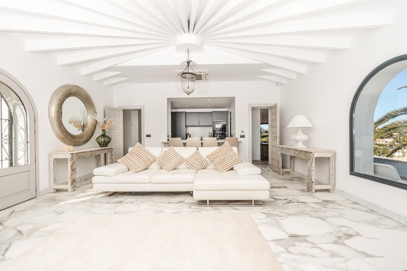 Circular living room featuring radial white ceiling beams, veined marble flooring, and large arched windows overlooking the Mediterranean landscape.