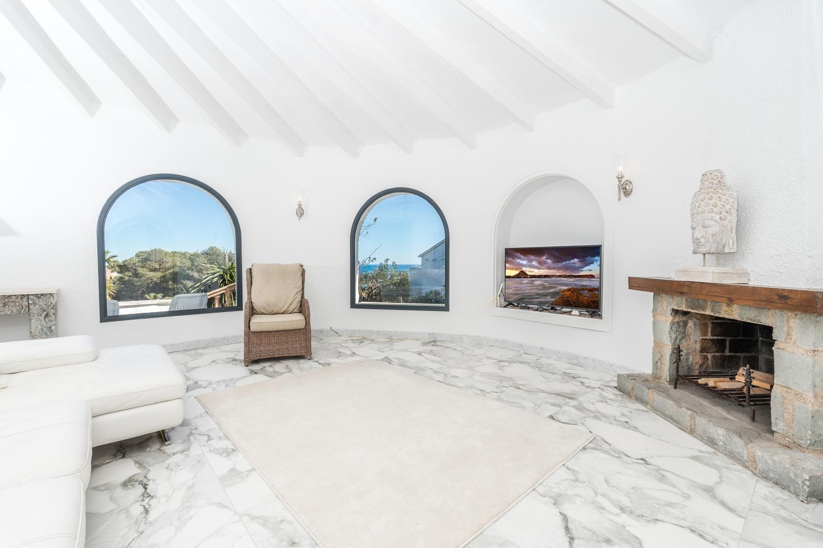 Circular living room featuring white beamed ceilings, marble flooring, and a stone fireplace. Arched windows provide natural light and views of the Mediterranean landscape.