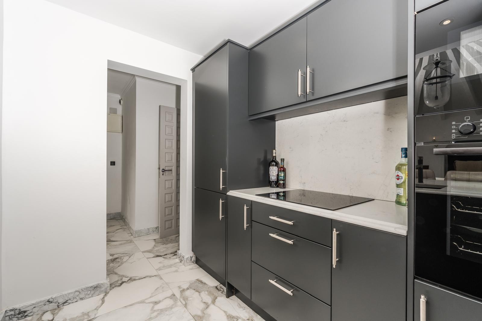 Contemporary kitchen featuring dark grey cabinetry, white marble countertops, and an integrated induction hob over elegant marble-veined flooring.
