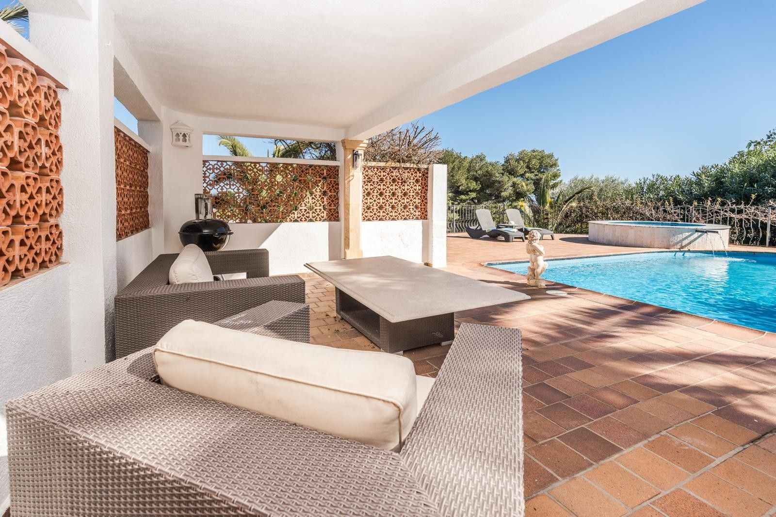 Covered porch featuring grey rattan lounge seating and terracotta lattice walls, overlooking the swimming pool and circular outdoor jacuzzi.