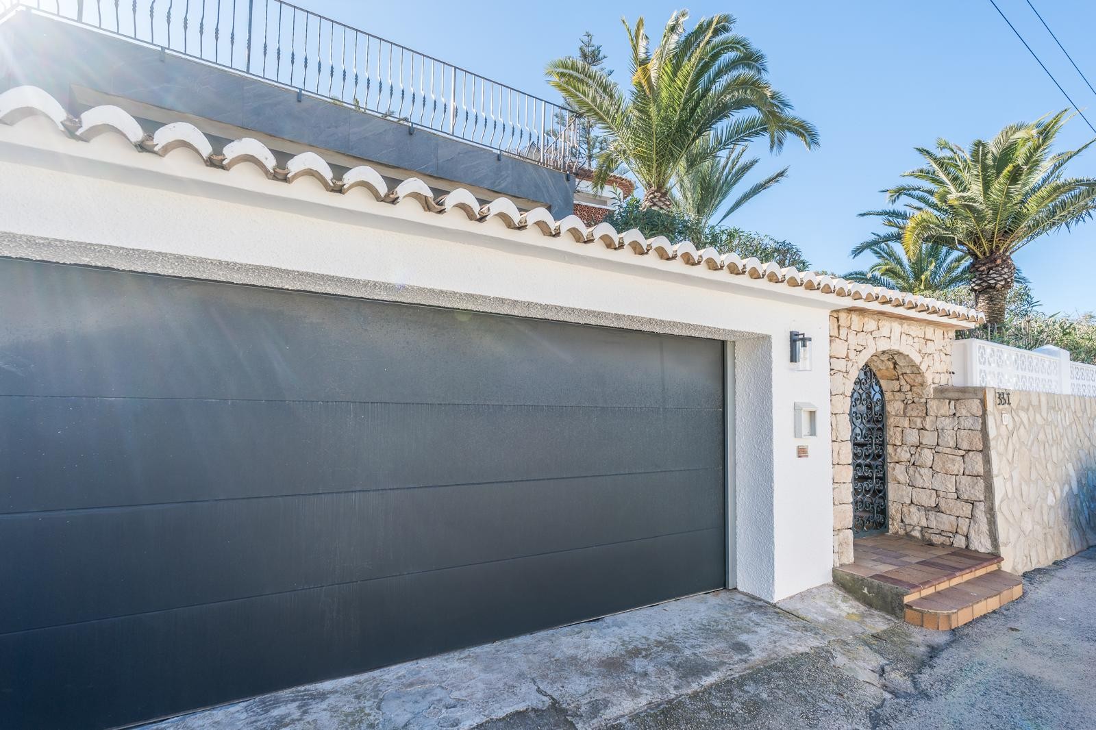 Exterior view featuring a dark grey double garage door, a traditional stone archway with a wrought-iron gate, and a natural stone wall lined with palm trees.