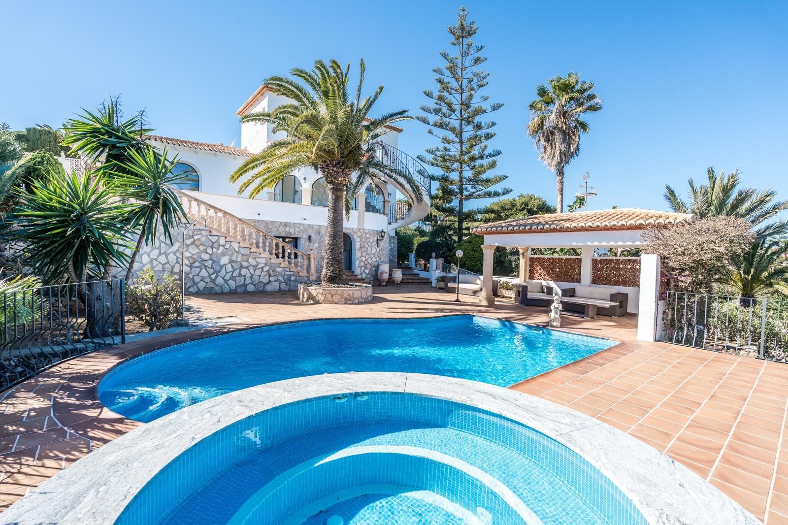 Exterior view featuring a renovated swimming pool, circular jacuzzi, and terracotta terrace, complemented by a Mediterranean garden with palms and a shaded lounge.