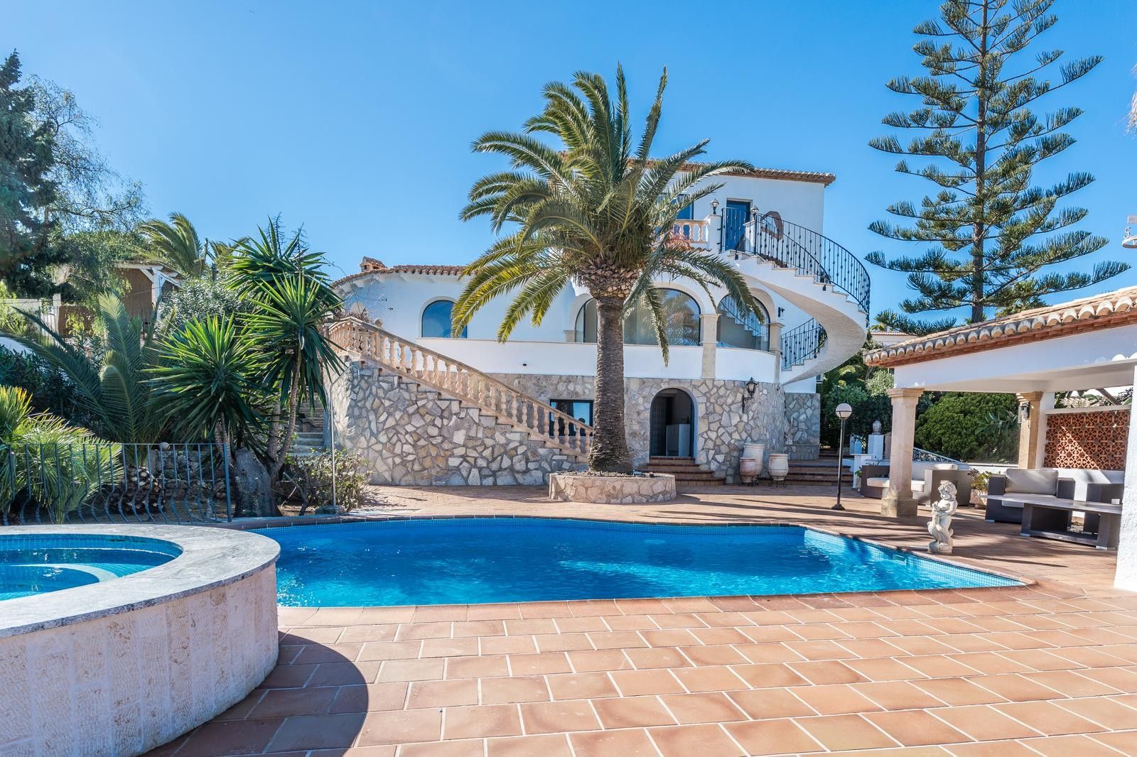 Exterior view of the villa featuring a renovated pool, circular jacuzzi, and stone masonry. The scene includes a spiral staircase and a Mediterranean garden with palms.