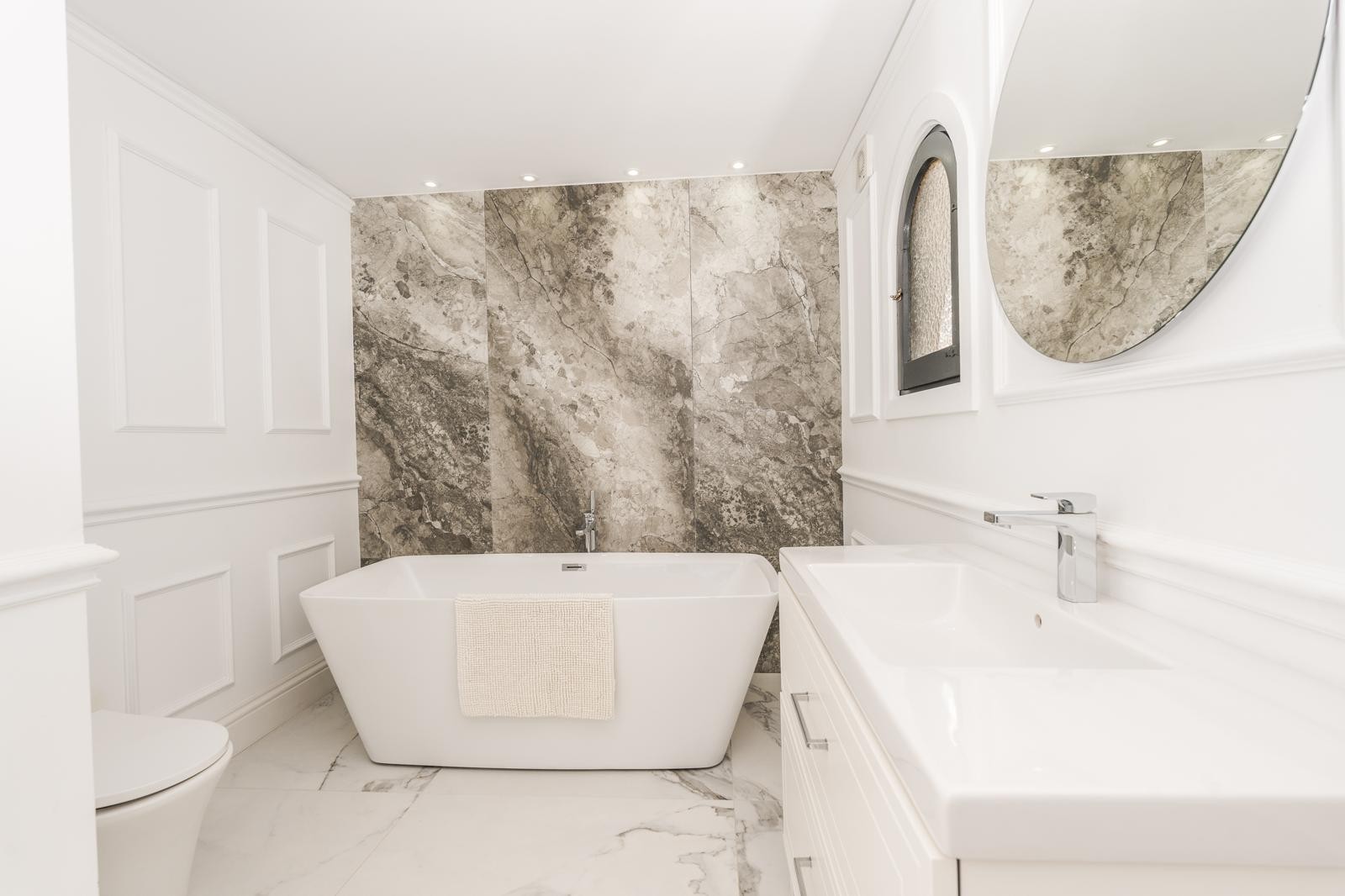 Master suite bathroom featuring a freestanding bathtub, marble-veined accent wall, and classic wall paneling. Includes a white vanity, round mirror, and recessed lighting.