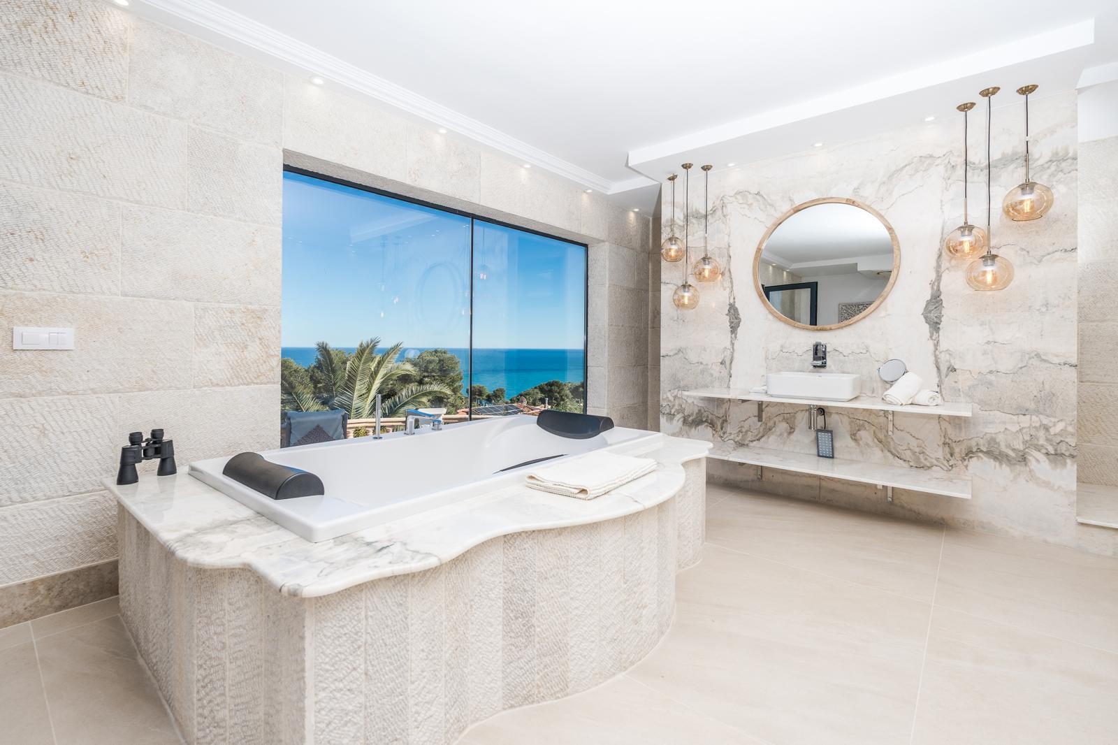 Master suite bathroom featuring a marble-encased bathtub with sea views, natural stone walls, and a vanity area with book-matched marble and glass pendant lighting.