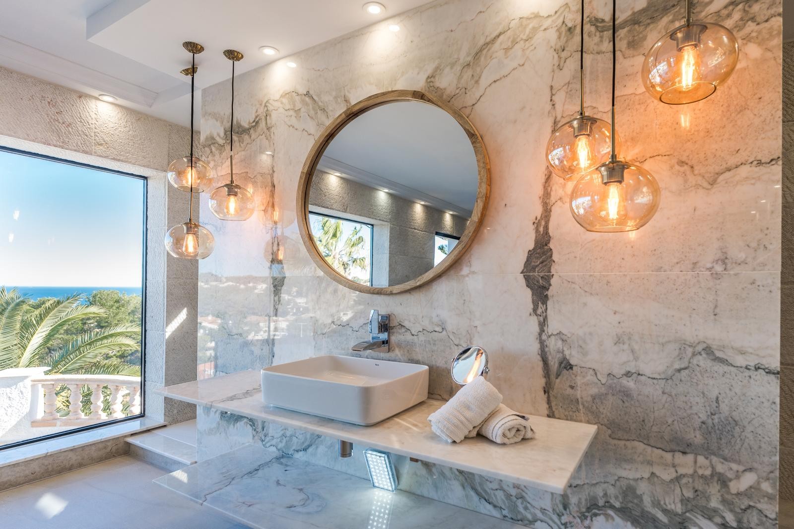 Master suite bathroom featuring book-matched marble walls, a vessel sink, circular wooden mirror, and designer pendant lighting with coastal Mediterranean views.