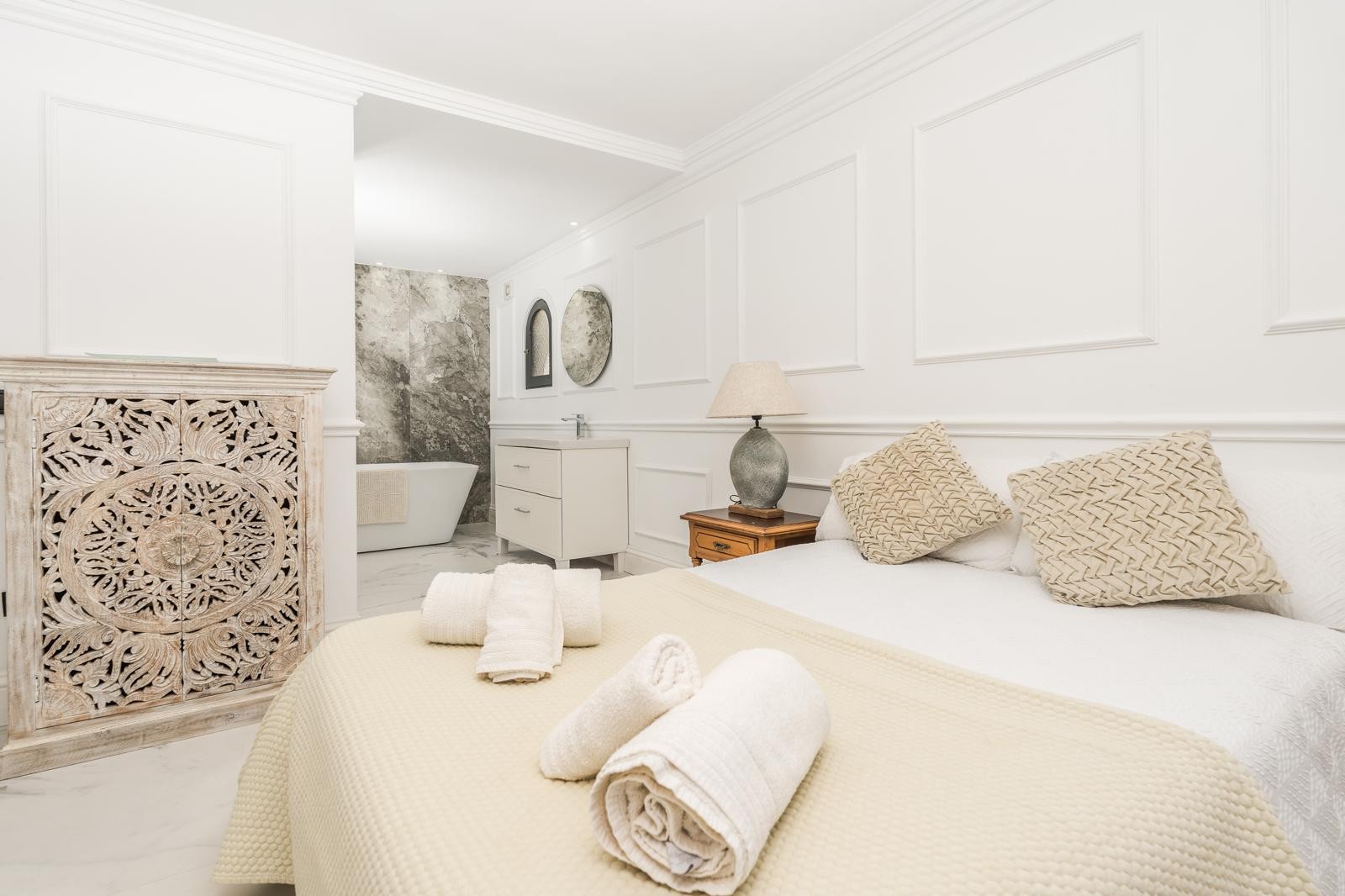 Master suite featuring decorative wall moldings, a hand-carved wooden cabinet, and an open-concept bathroom with a freestanding tub and marble walls.