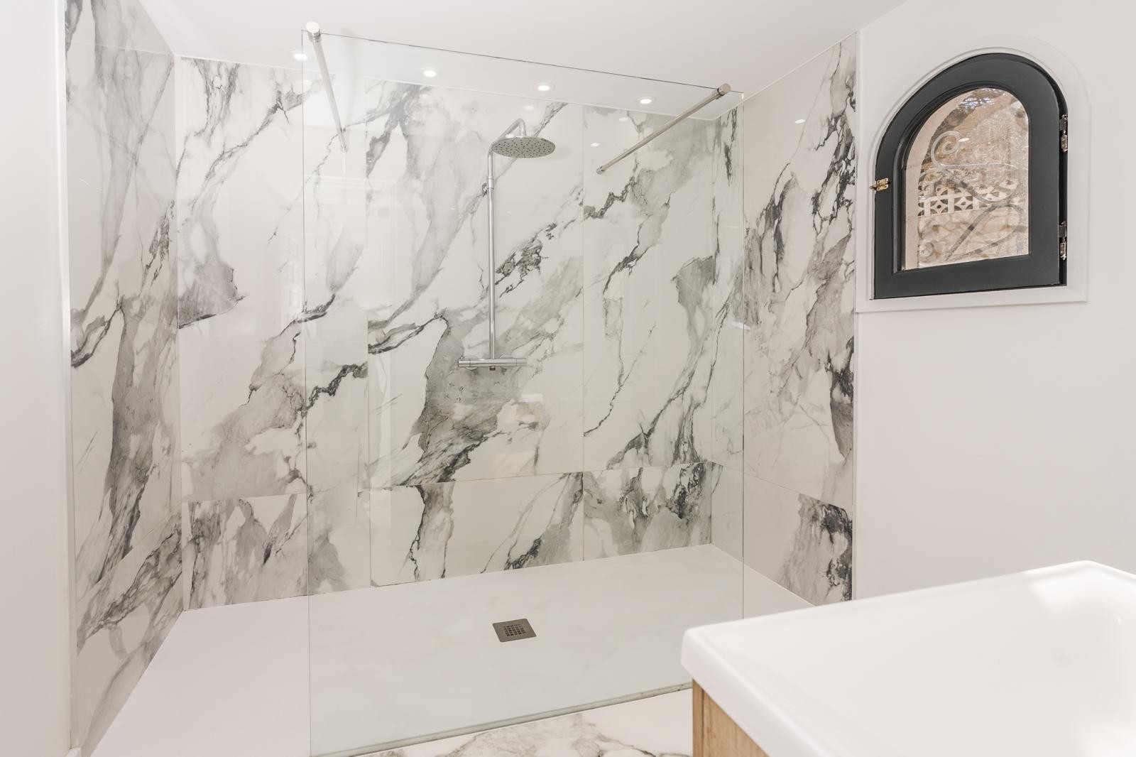 Modern walk-in shower featuring large-format marble tiles, a fixed glass screen, chrome rain showerhead, and a traditional arched window.