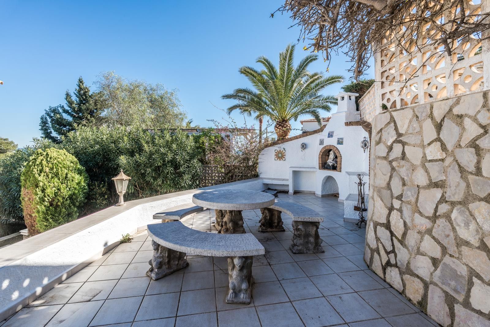 Outdoor terrace featuring a carved stone table and benches, a built-in white masonry barbecue, and natural stone walls amidst Mediterranean greenery.