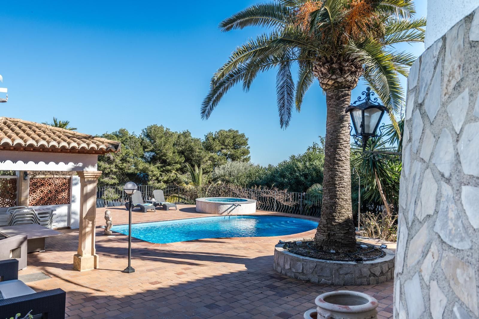 Outdoor terrace featuring a curved swimming pool, circular hot tub, and a mature palm tree. The area includes a covered porch with stone pillars and classic terracotta tiling.