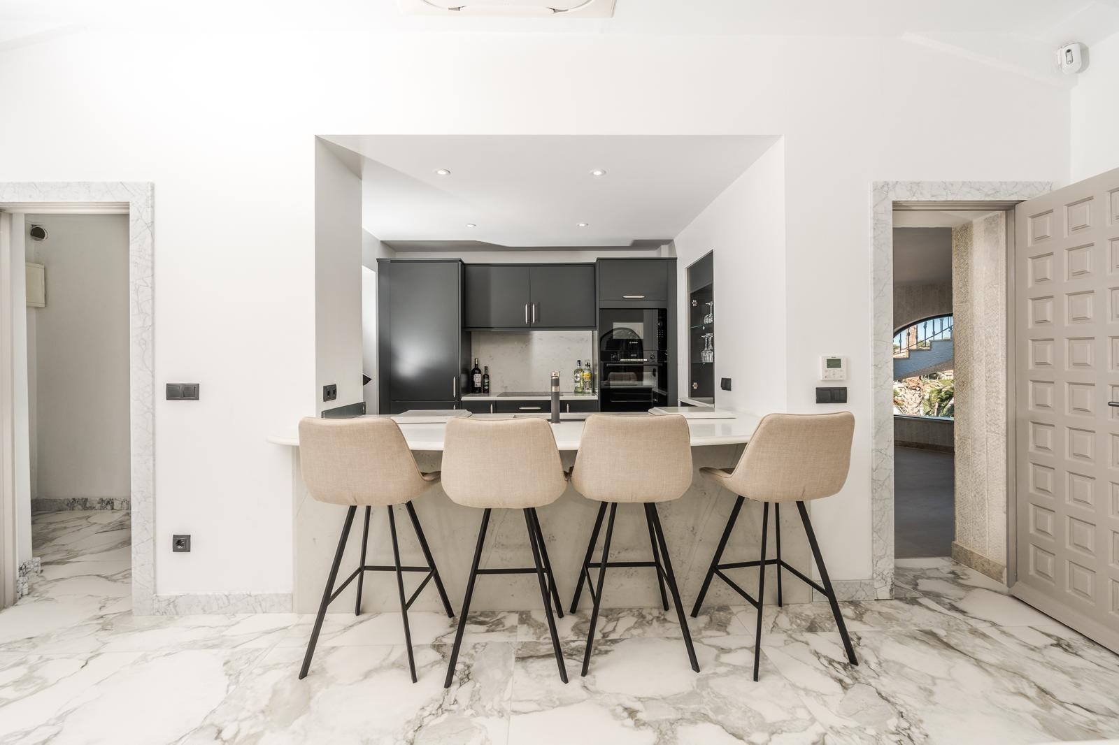 Semi-open kitchen featuring dark cabinetry, marble countertops, and a breakfast bar with upholstered stools on elegant marble-effect flooring.