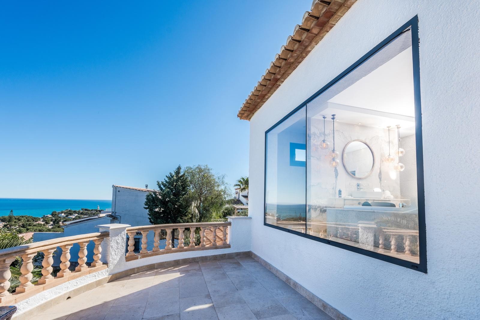 Upper terrace featuring a stone balustrade and sea views, adjacent to a large window revealing a modern bathroom with marble walls and designer lighting.