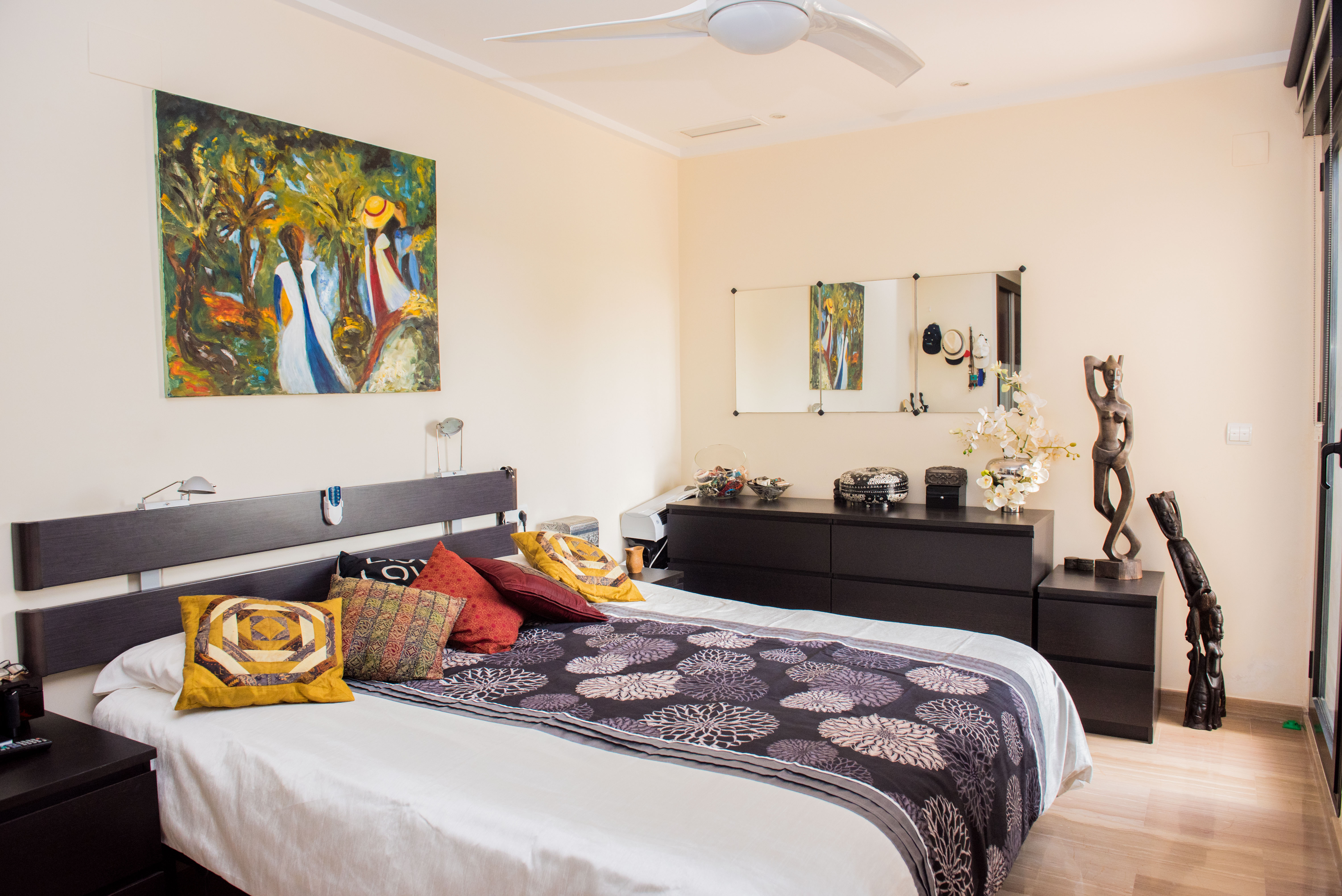 Bedroom featuring a dark wood headboard, matching dresser, and white ceiling fan. Includes vibrant wall art, decorative sculptures, and light-toned flooring.