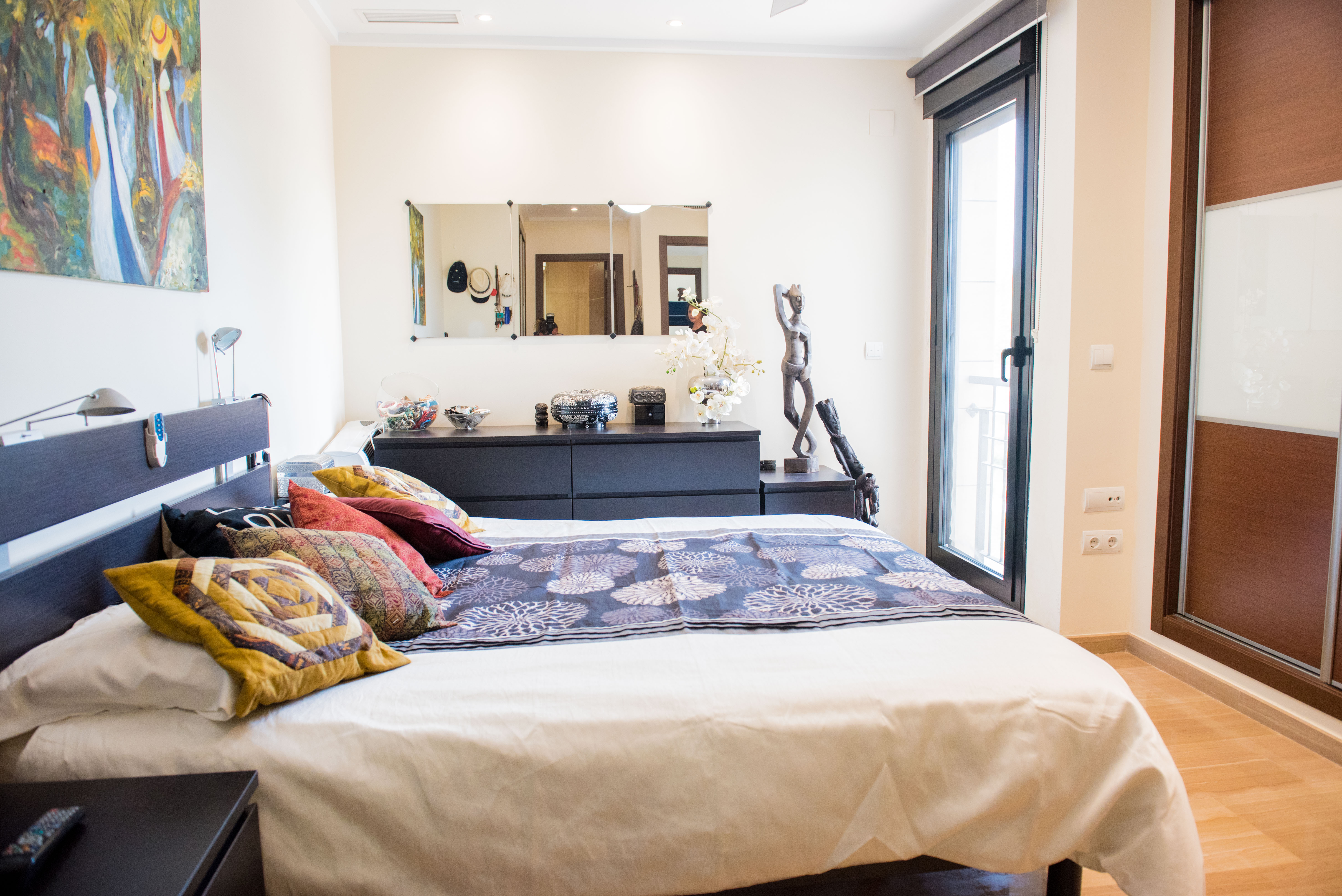 Bedroom with dark wood furniture, built-in sliding wardrobes, and a glass door for natural light. The space includes a dresser with a mirror and light wood flooring.
