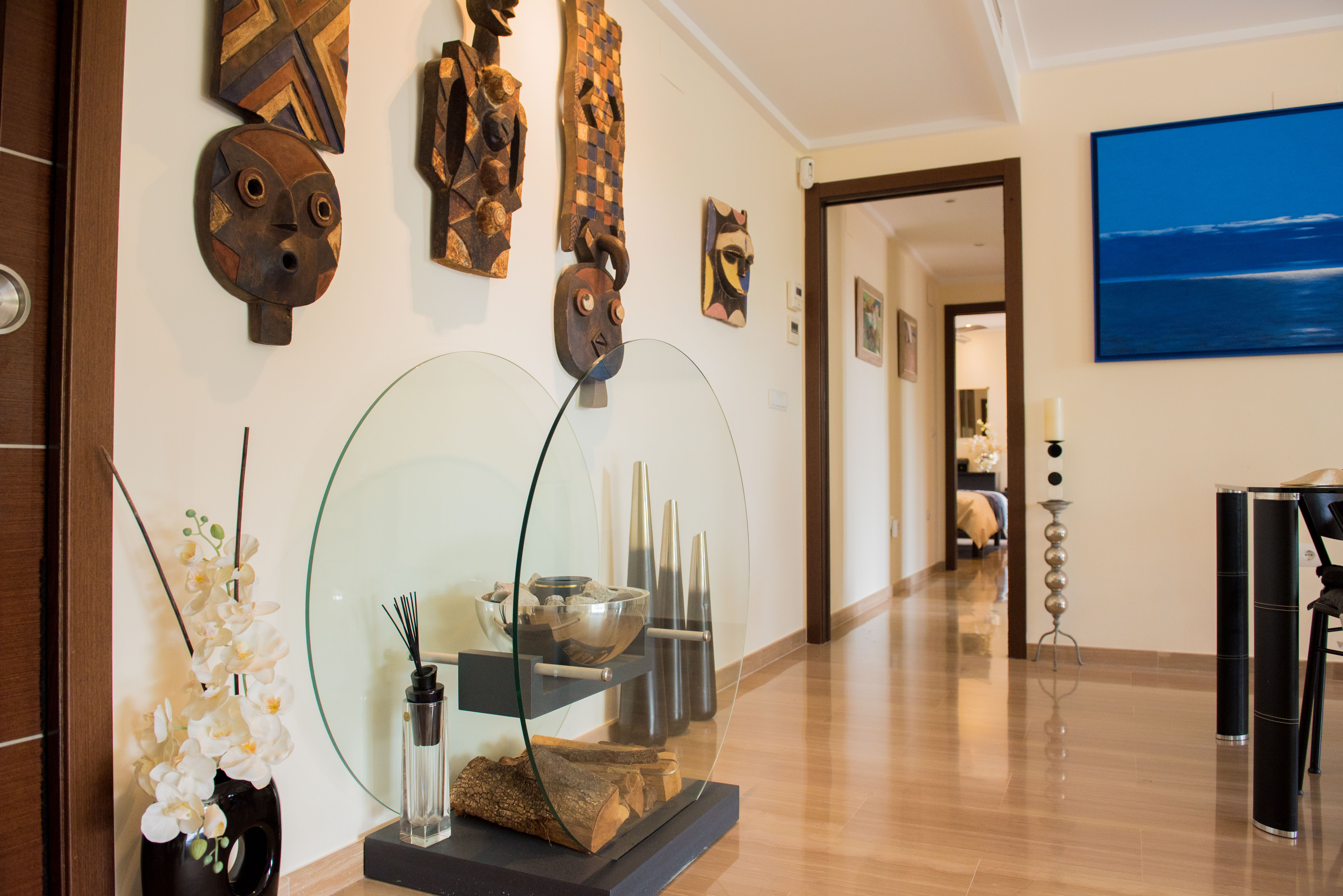 Bright hallway with glossy wood-effect tiled floor, adorned with tribal wooden masks and a contemporary glass display of decorative logs and orchids. A blue abstract artwork is visible.