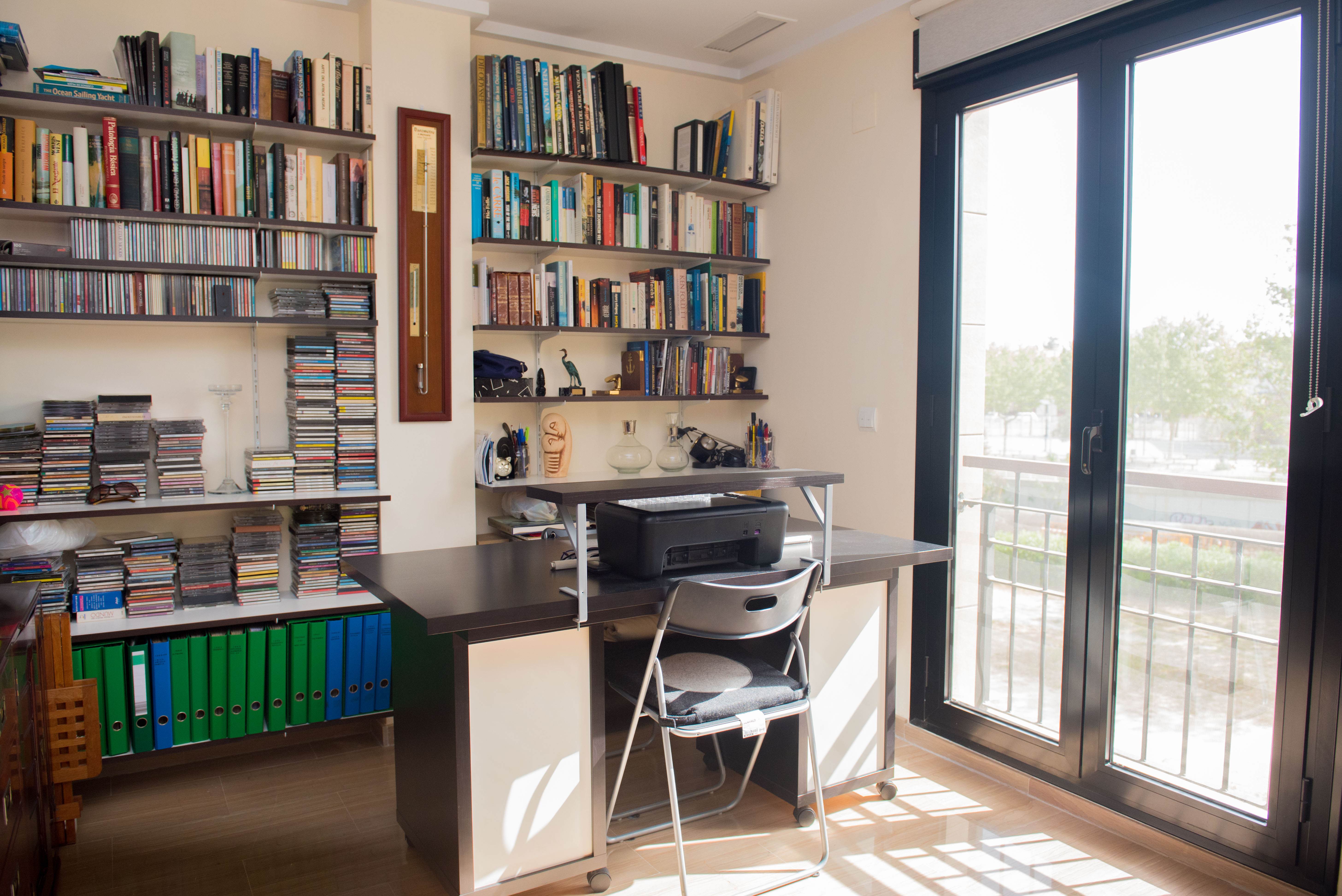 Bright home office featuring a dark wood desk, extensive bookshelves filled with books and CDs, and direct access to an outdoor balcony via a glass door.