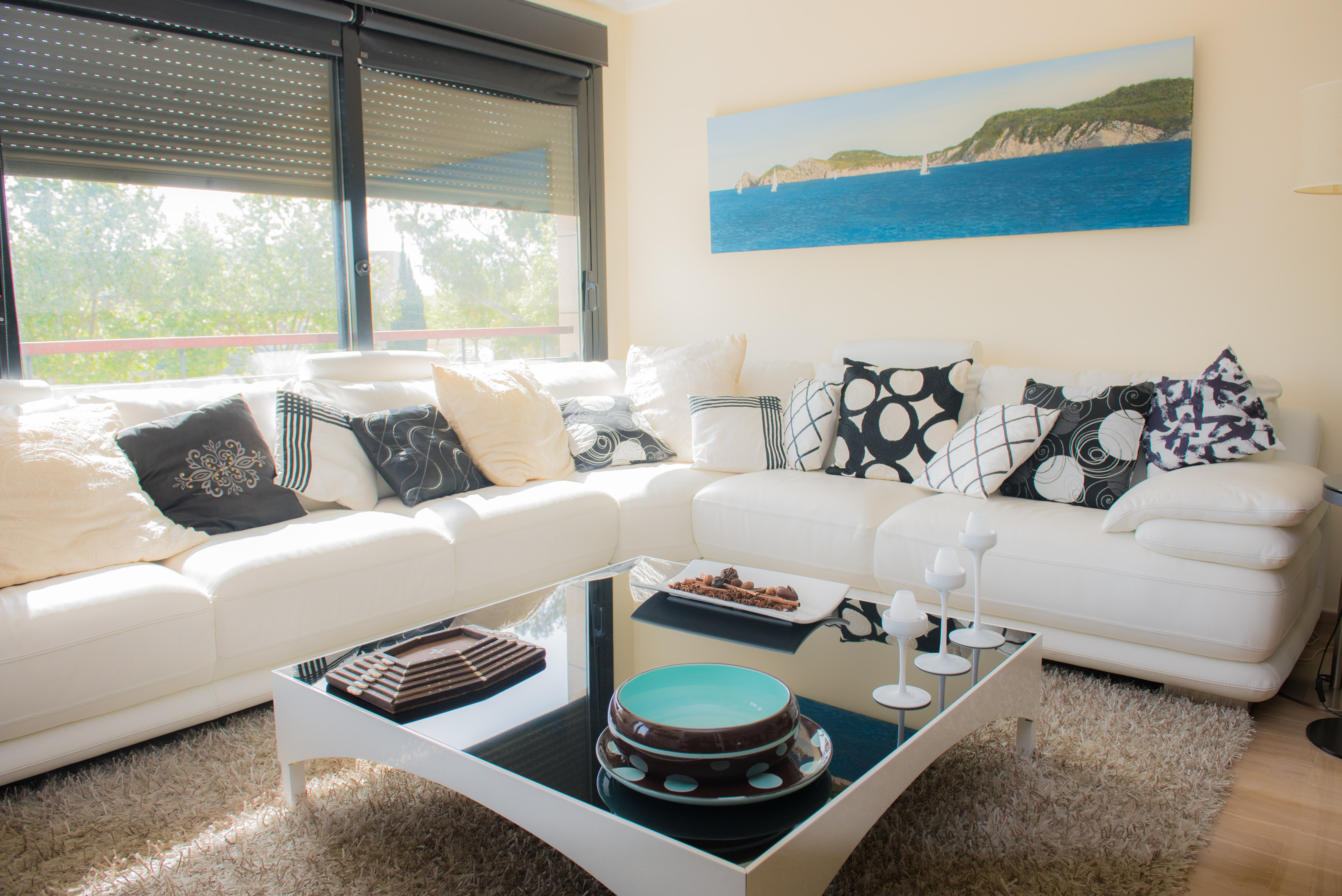 Bright living area featuring a white leather sectional sofa with patterned cushions and a black glass coffee table. Natural light enters through large windows with shutters.