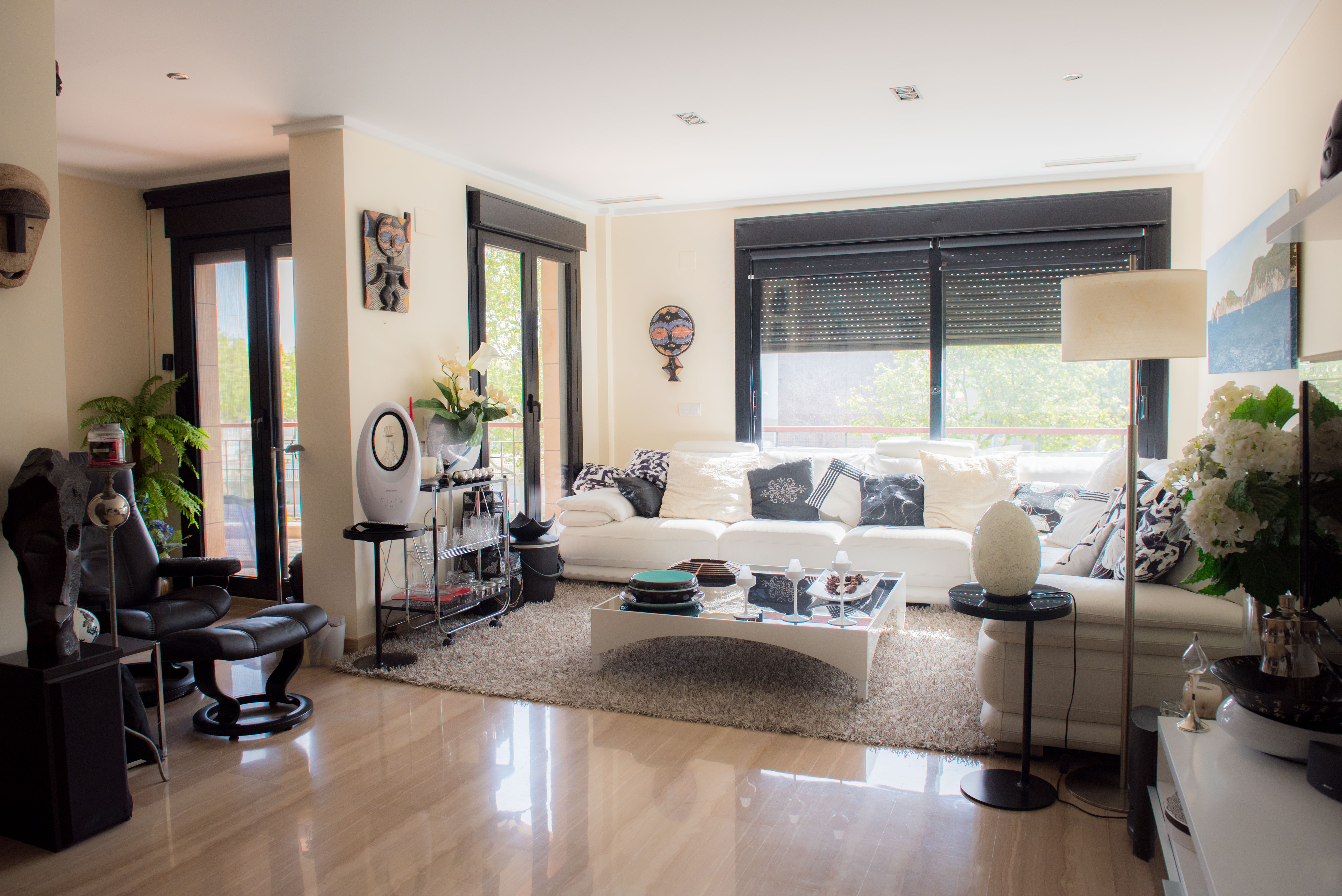 Bright living area featuring polished stone flooring, a large white sectional sofa, and floor-to-ceiling windows with integrated shutters.