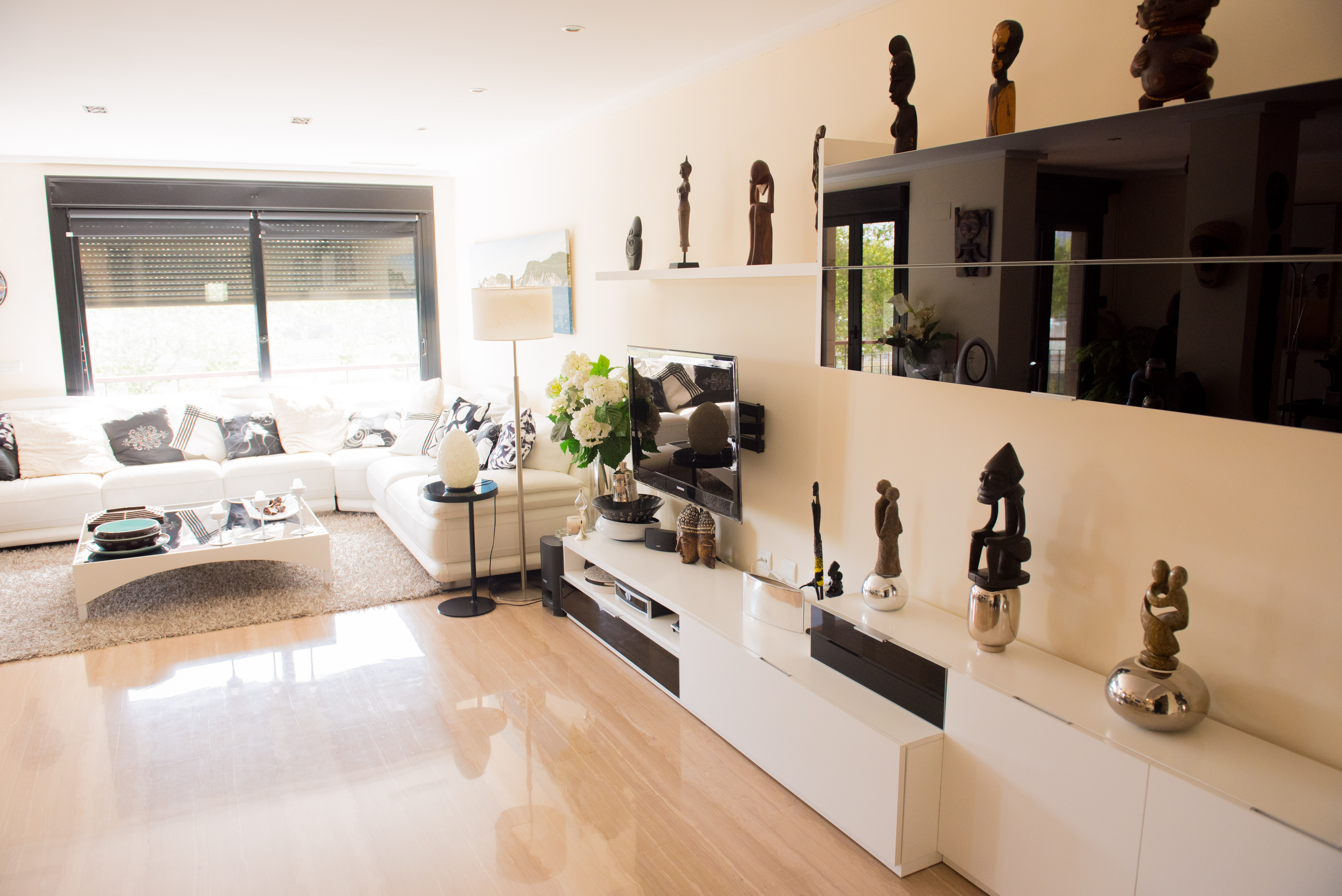 Bright living room featuring a white sectional sofa, polished light-toned tile flooring, and a modern media console. Natural light enters through the window with roller blinds.