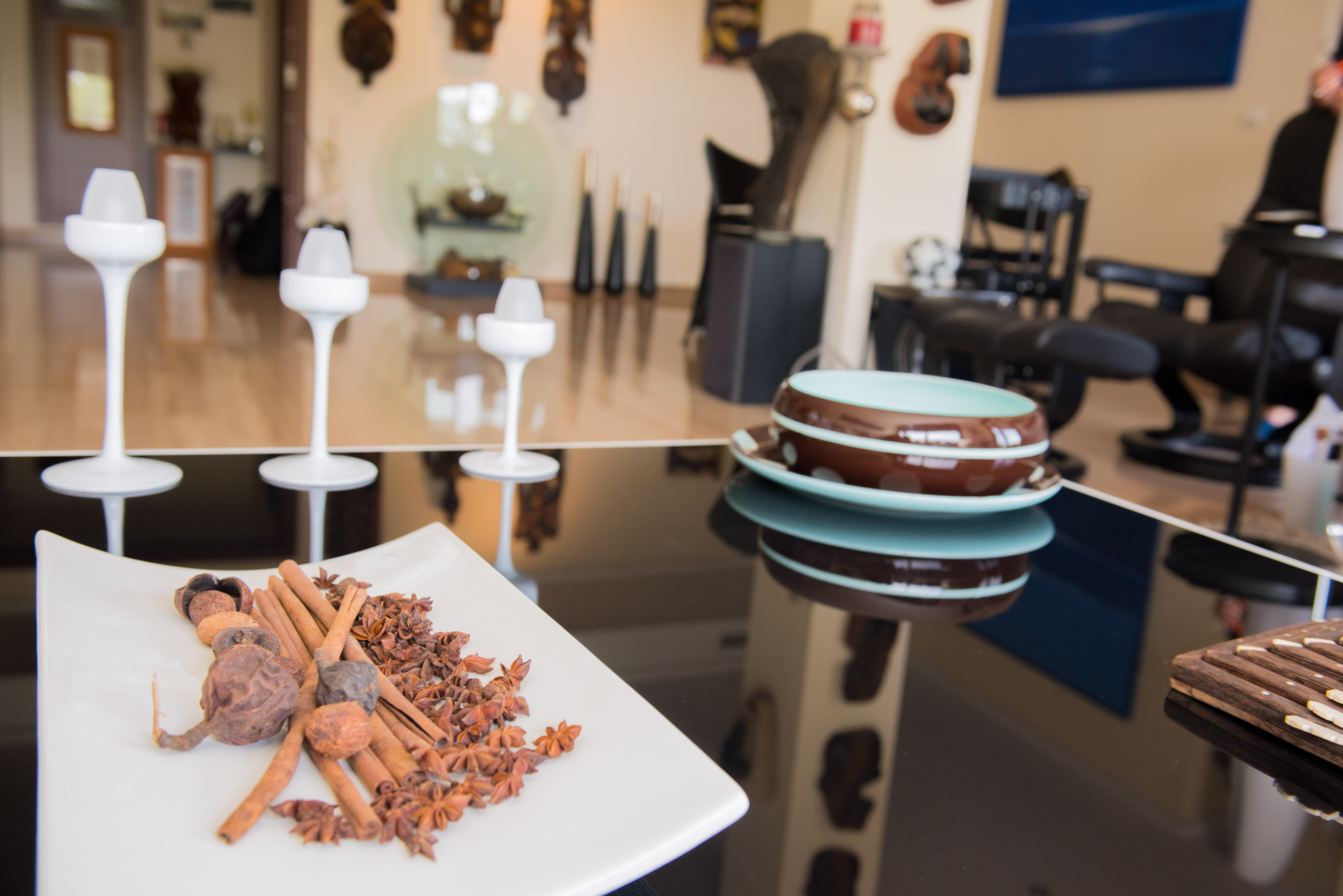 Close-up of a reflective table featuring a plate with cinnamon sticks and star anise, modern white candle holders, and ceramic bowls. The background reveals a decorated interior.