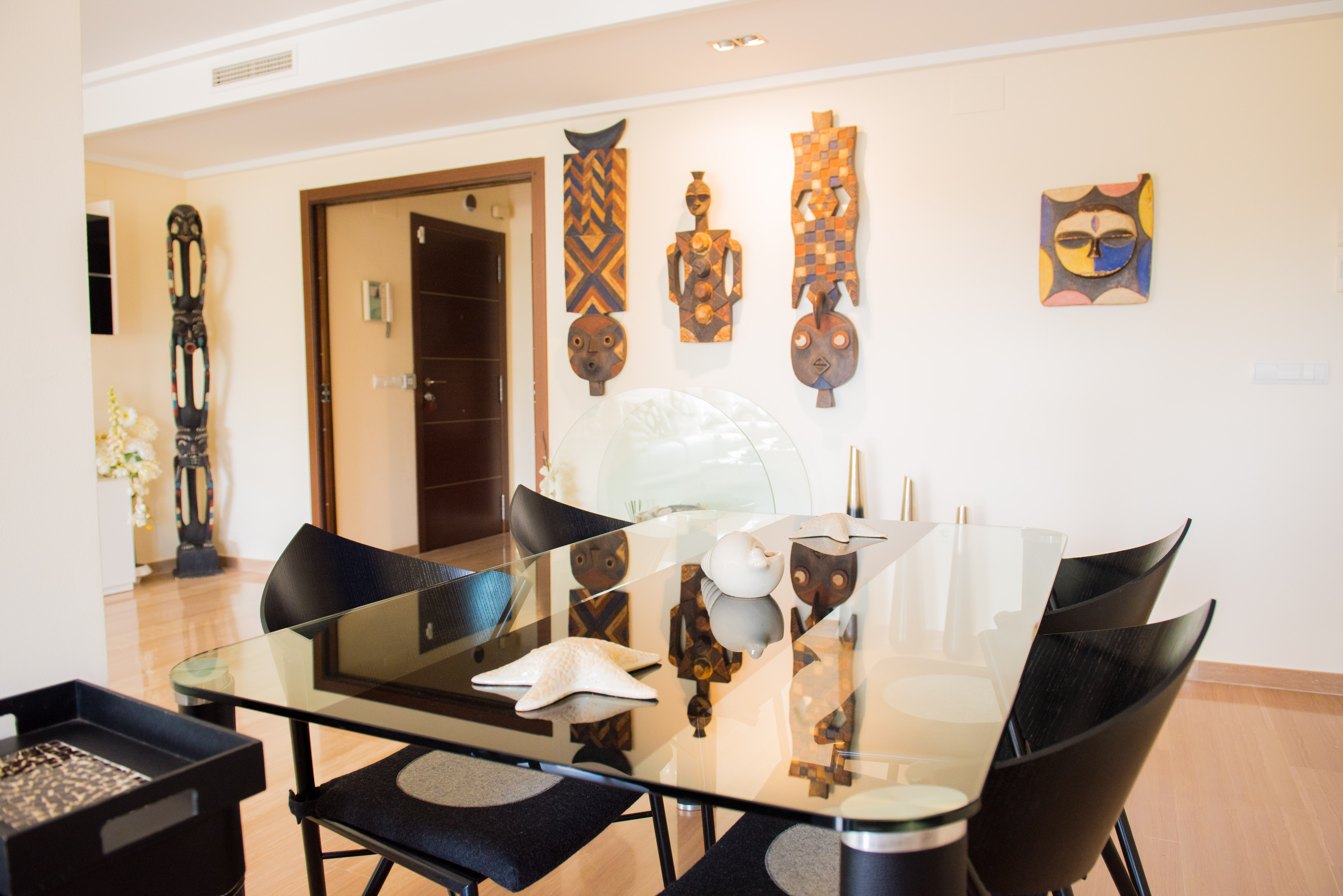Contemporary dining area featuring a glass table, sleek black chairs, and light wooden flooring. Unique ethnic masks adorn the wall.