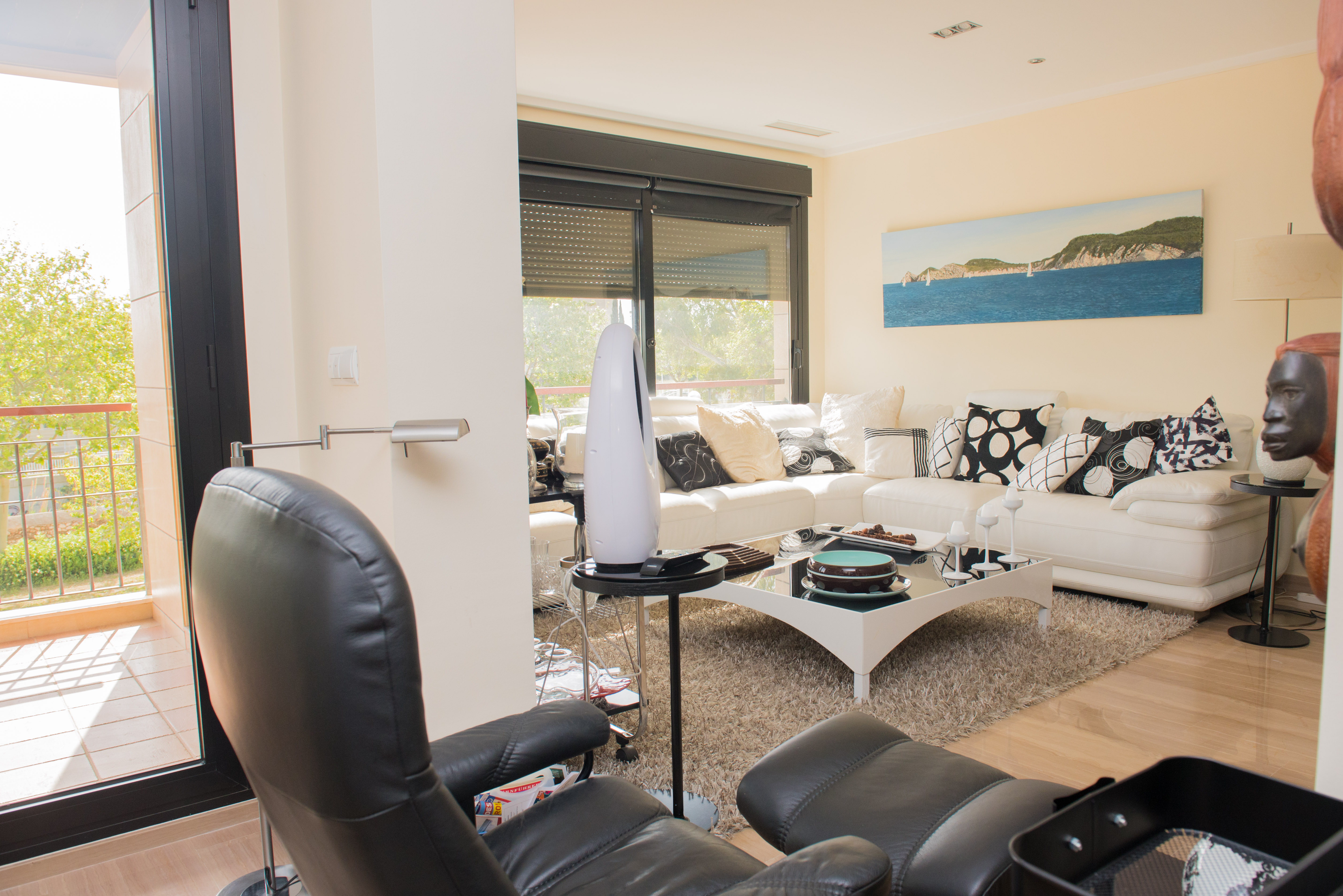 Contemporary living room featuring a white sectional sofa, black glass coffee table, and light wood-look tiled floor. Direct access to a terrace with metal railings and green tree views.