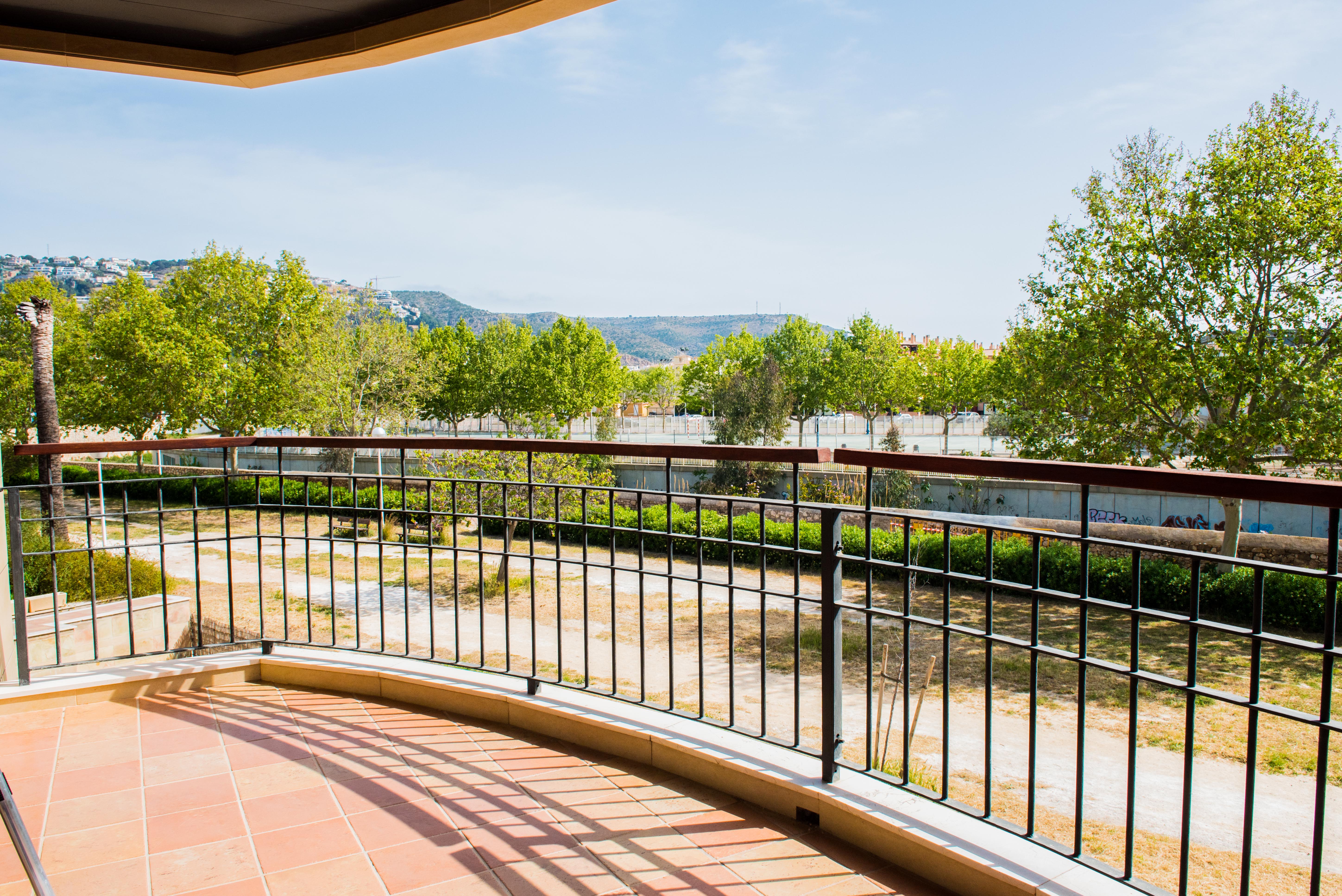 Covered terrace featuring terracotta tile flooring and a wrought iron railing with a wooden handrail, overlooking a green park and distant mountains.