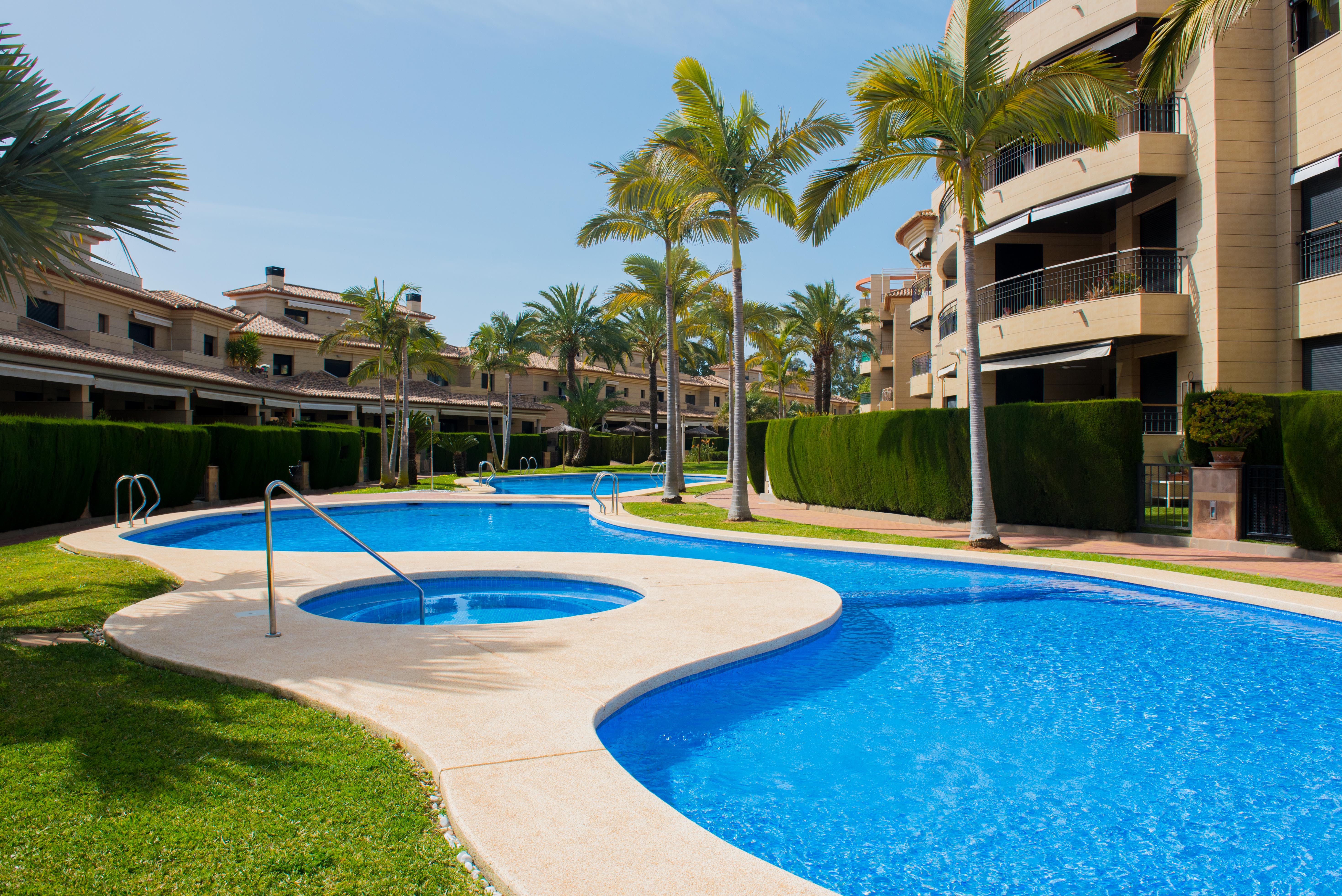 Curved communal swimming pool with integrated whirlpool, surrounded by manicured lawns, palm trees, and modern apartment buildings with stone facades.