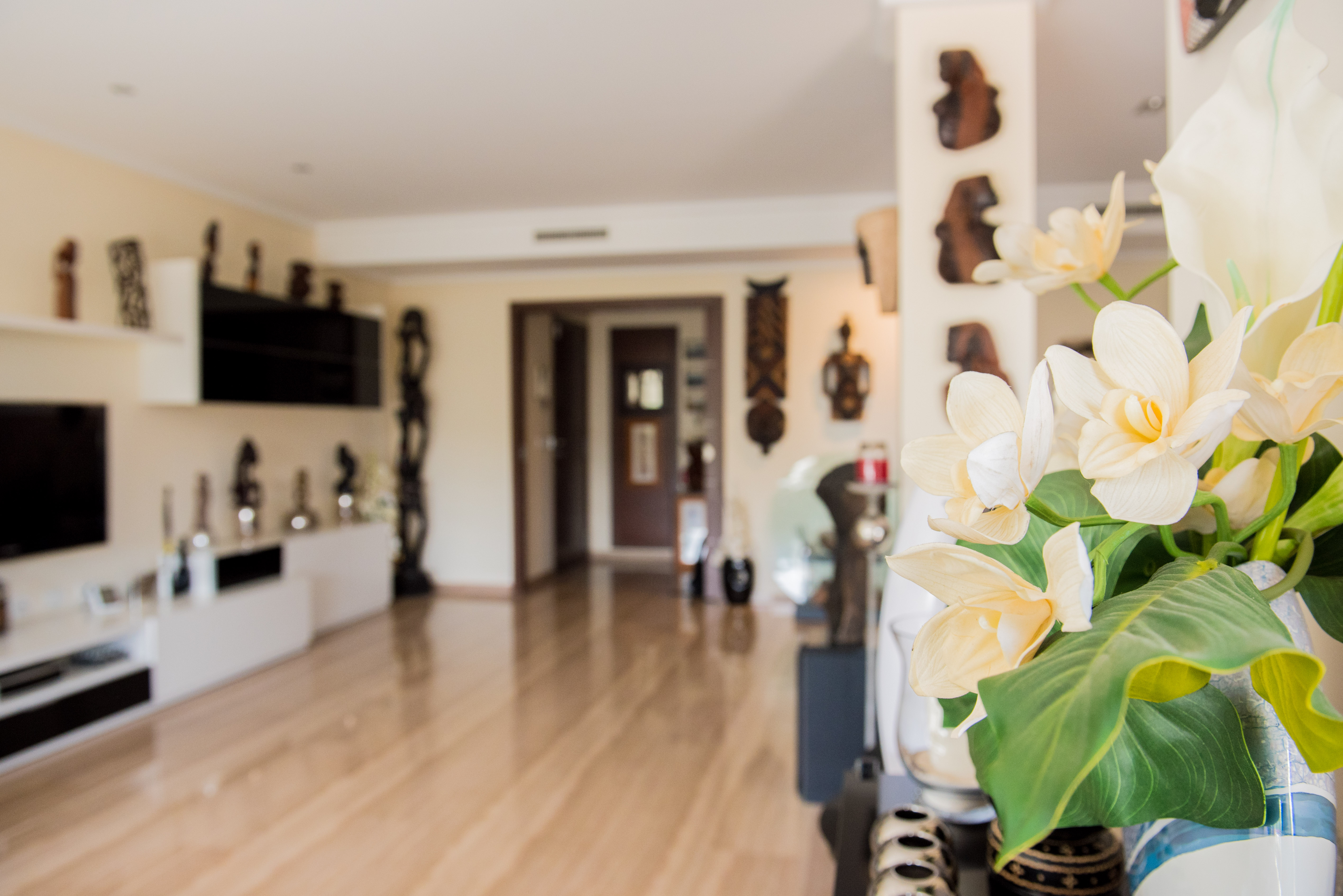Foreground vase with elegant white and yellow artificial flowers. Bright interior with light wood flooring, leading to a modern living area with decorative wall art.