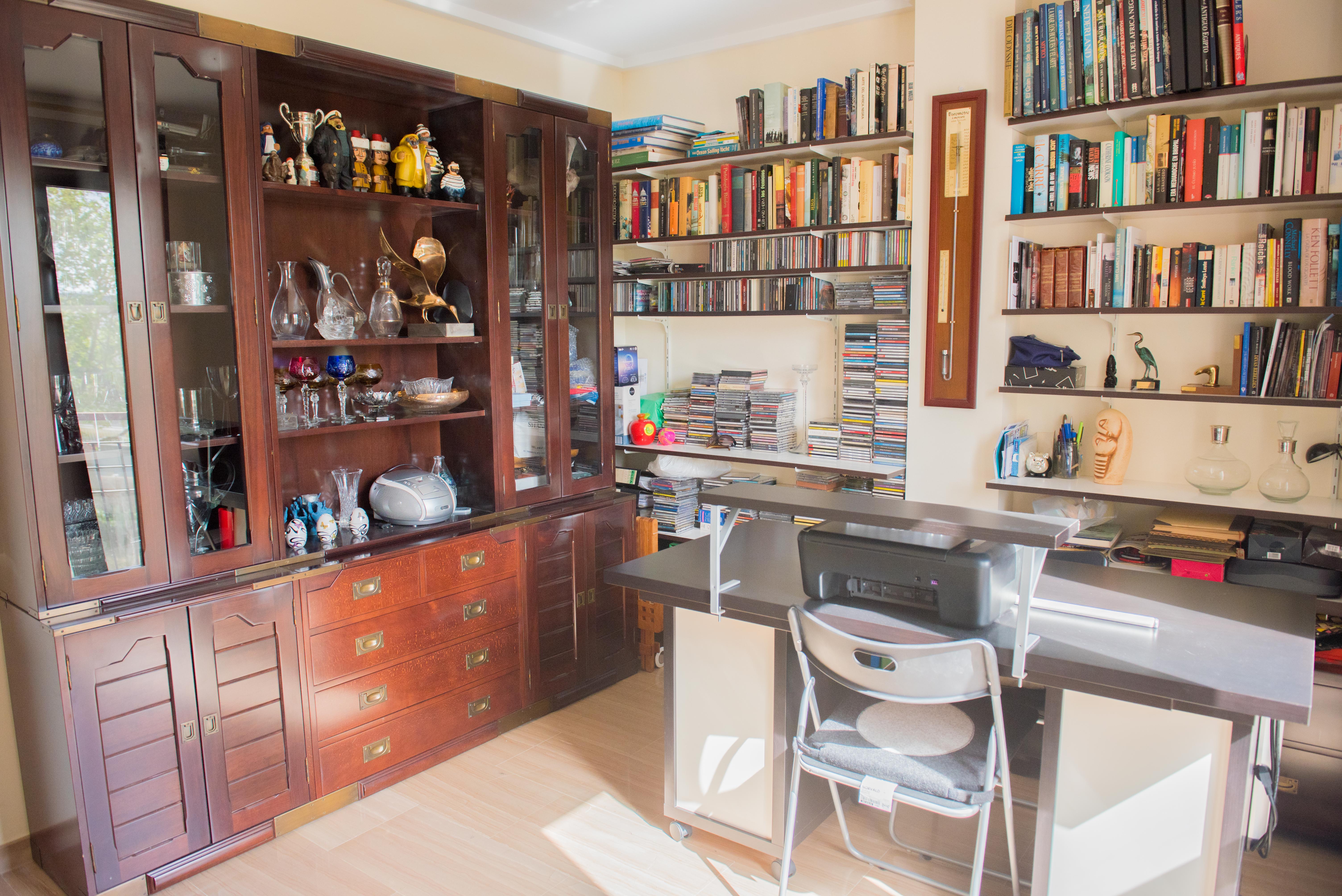 Home office featuring a large dark wood display cabinet, extensive wall-mounted bookshelves, and a functional desk area with natural light and tiled flooring.