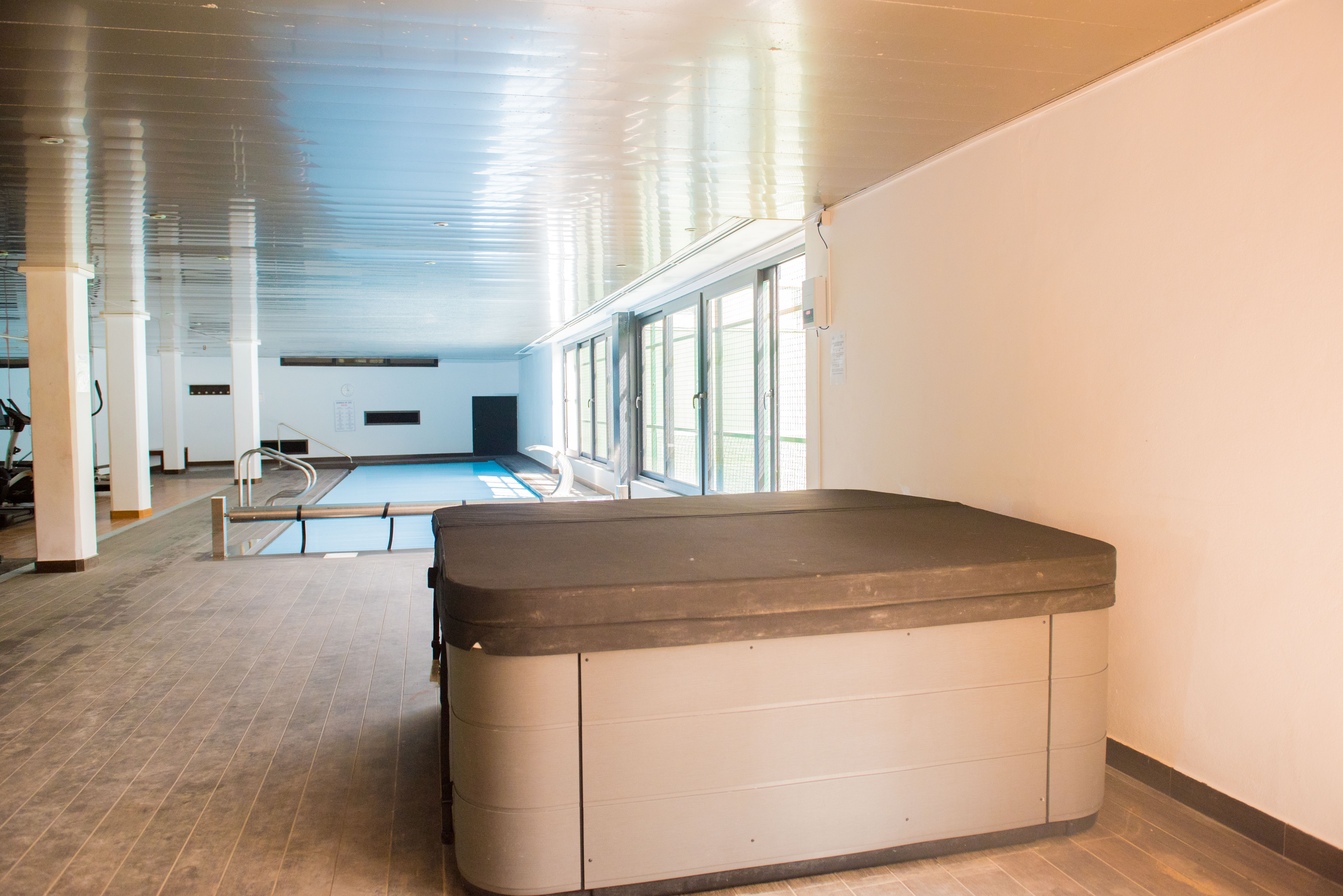 Indoor wellness area featuring a covered hot tub, a swimming pool, and a gym zone with natural light from large side windows.