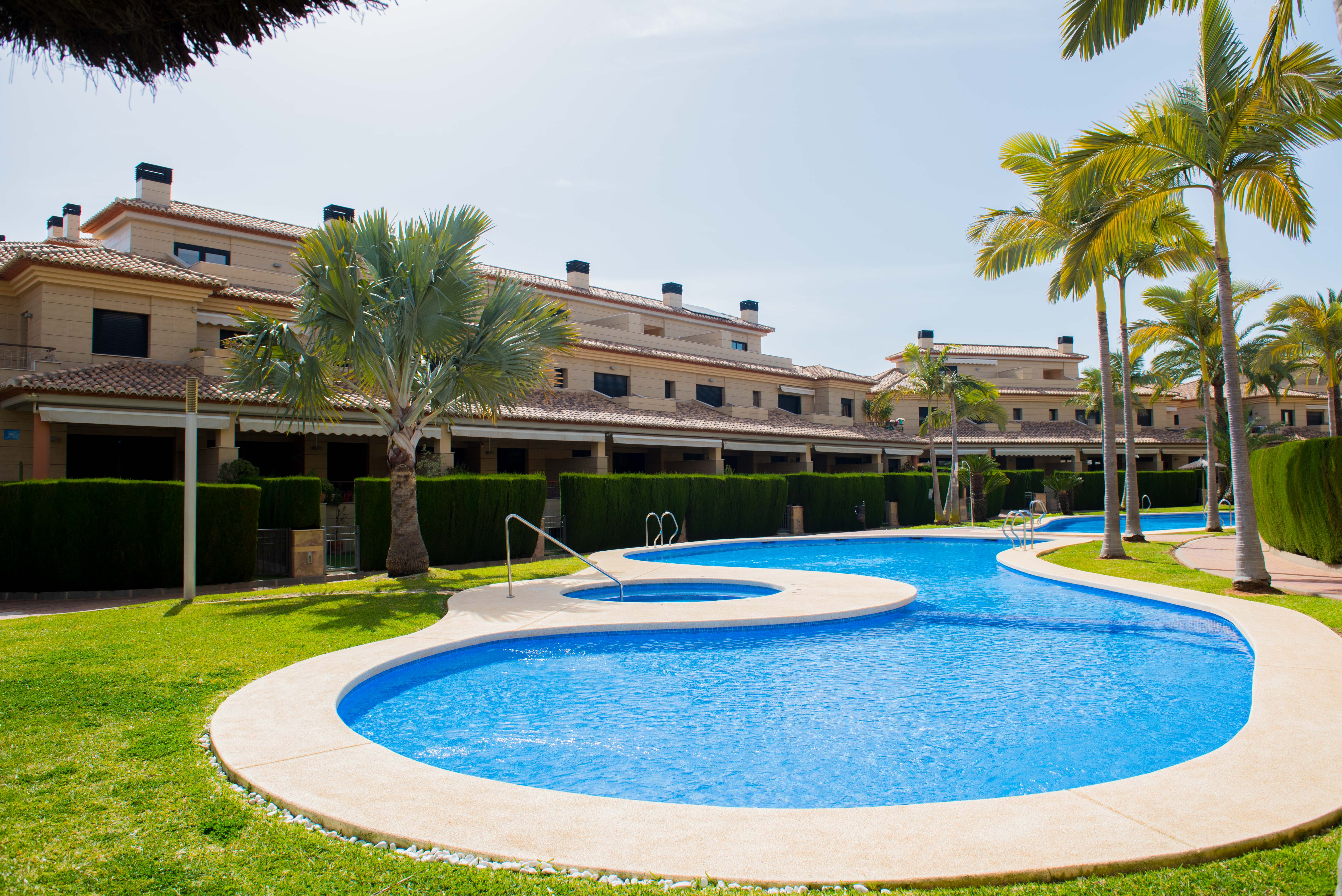 Large communal swimming pool with an integrated circular spa, surrounded by manicured lawns and palm trees adjacent to stone-cladded buildings.