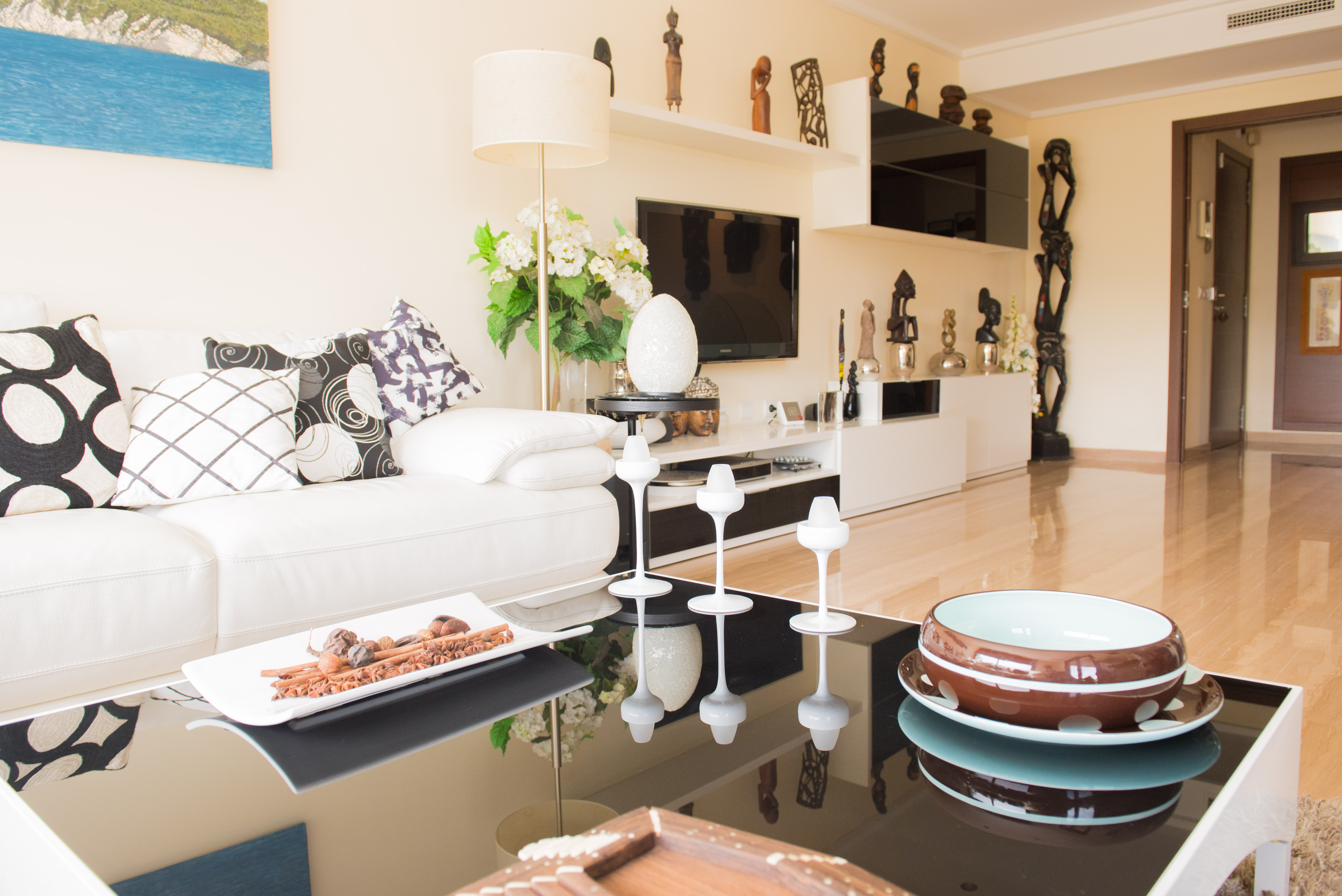 Living room featuring a white sofa, graphic cushions, and a glossy black coffee table with decorative accents. TV unit with shelves and African sculptures.