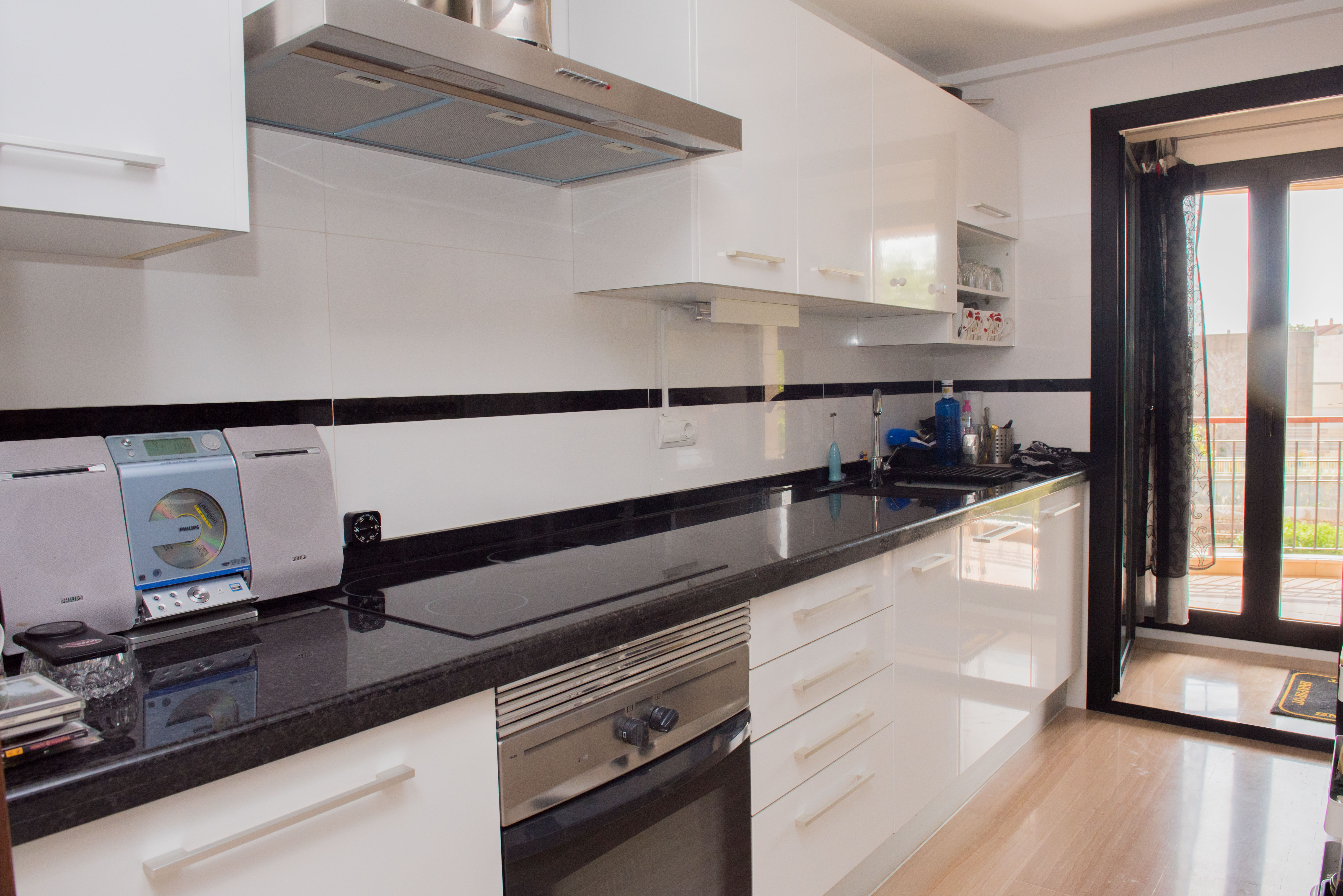 Modern kitchen featuring high-gloss white cabinetry, black granite countertops, and stainless steel appliances. A glass door leads to an outdoor balcony.