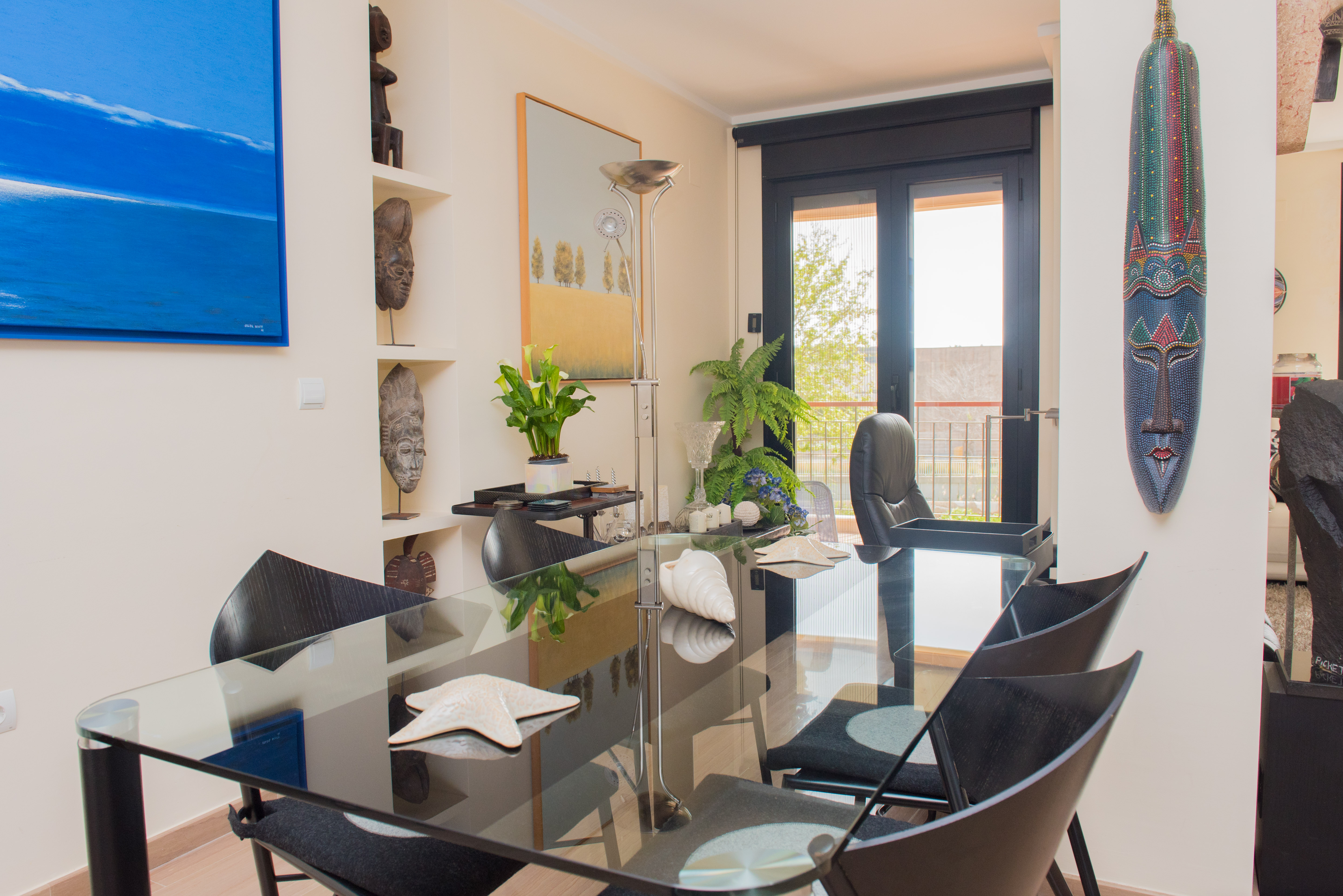 Spacious dining area featuring a glass table and black chairs, accented by a blue abstract painting and tribal masks on shelves. Large glass doors open to a balcony, providing ample natural light.