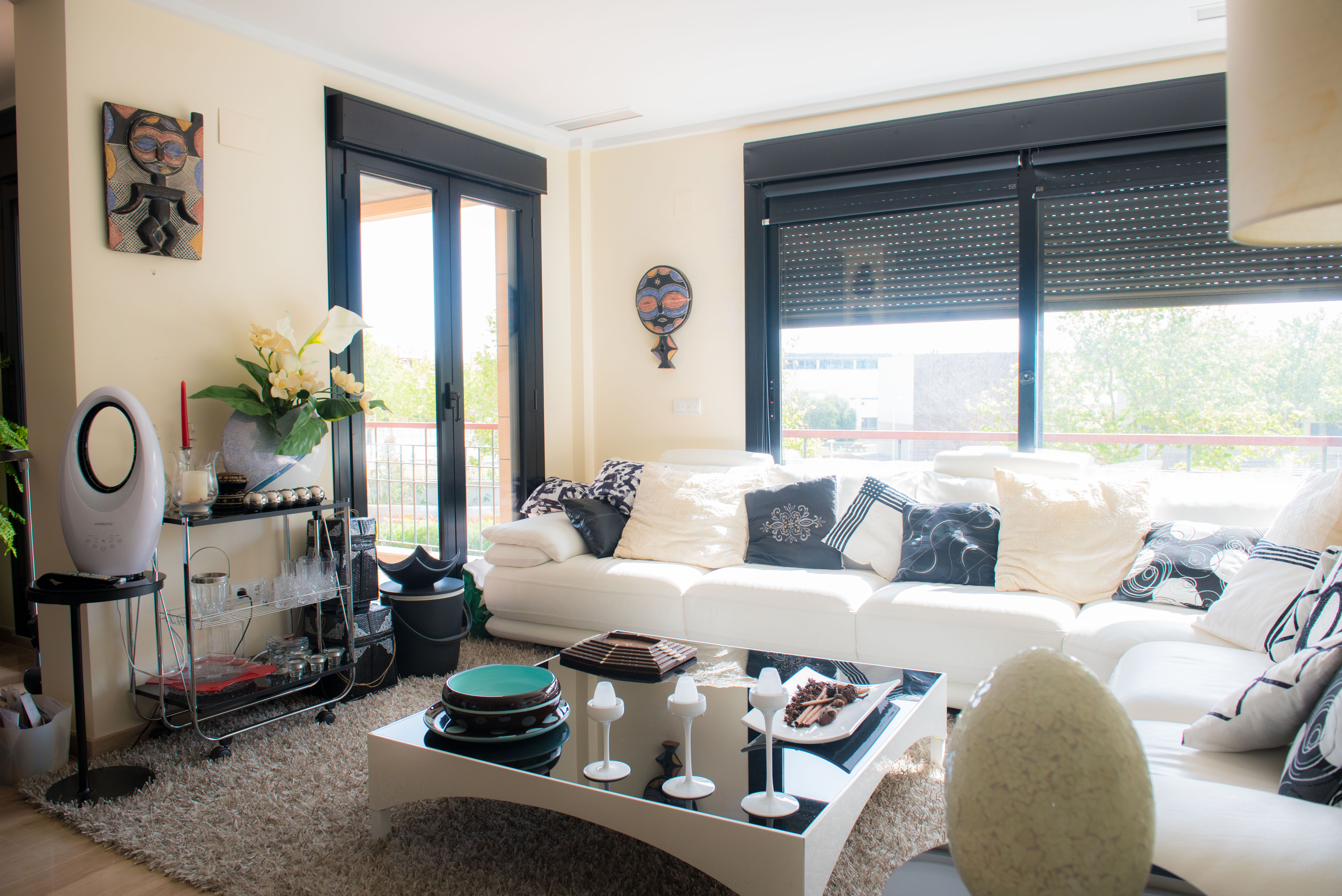 Spacious living room featuring a white sofa with diverse cushions, a plush rug, and a modern coffee table. Large windows with black frames offer views of outdoor greenery.