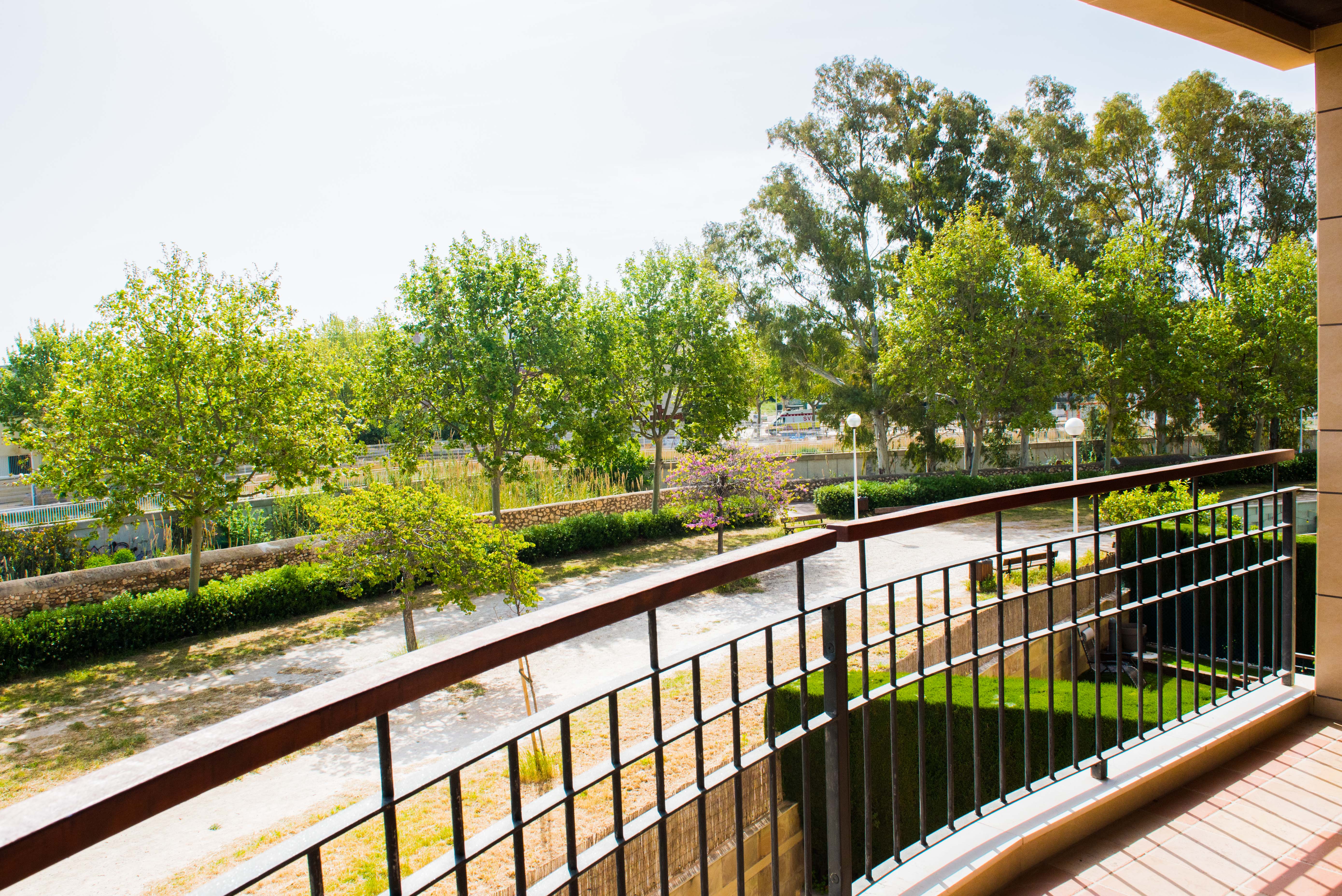 View from a private terrace showcasing lush communal gardens with mature trees, a winding path, and a stone wall, under a bright sky. The balcony features a classic metal railing and terracotta-style tiles.
