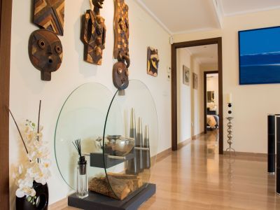 Bright hallway with glossy wood-effect tiled floor, adorned with tribal wooden masks and a contemporary glass display of decorative logs and orchids. A blue abstract artwork is visible.